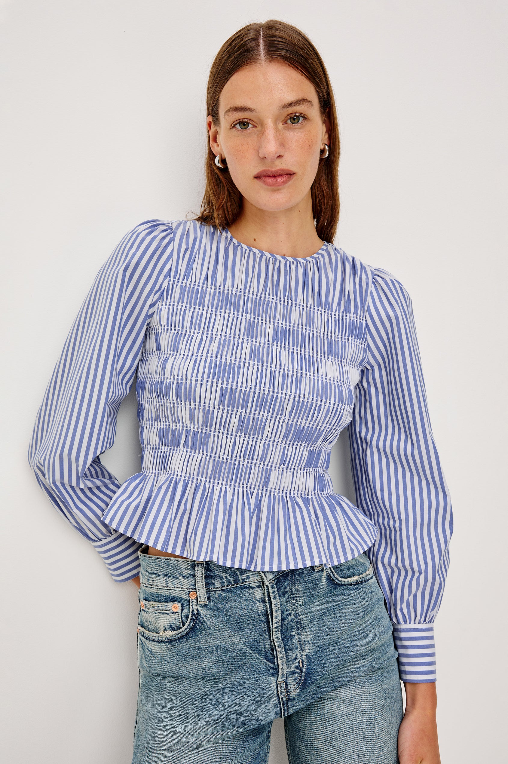 Tabi is a long sleeve crew neck top with puff sleeves and buttons at cuff, the bodice is pleated with a peplum hem in a blue Mariner White Stripe colorway. Front view featured on model.