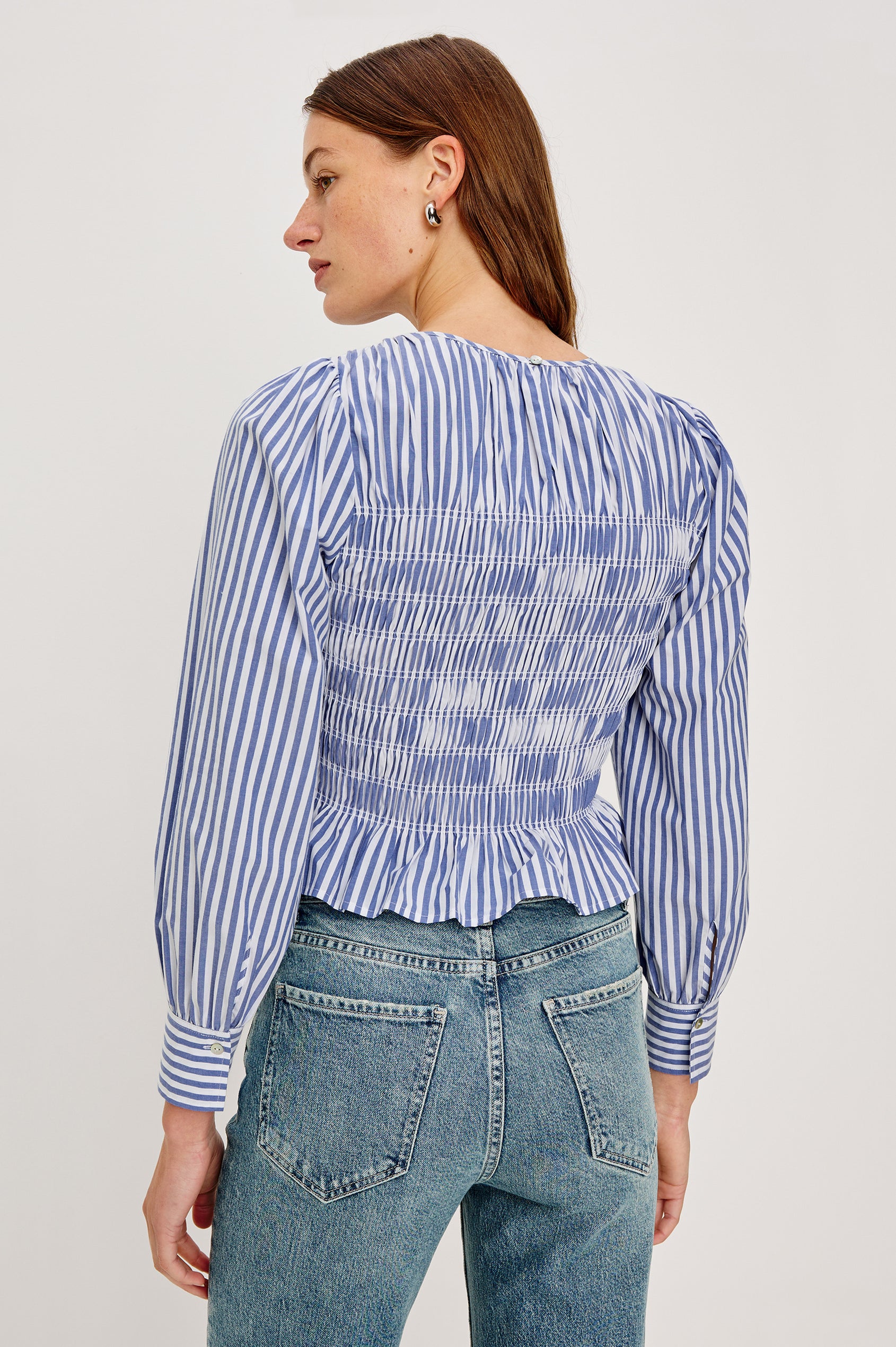 Tabi is a long sleeve crew neck top with puff sleeves and buttons at cuff, the bodice is pleated with a peplum hem in a blue Mariner White Stripe colorway. Back view featured on model.