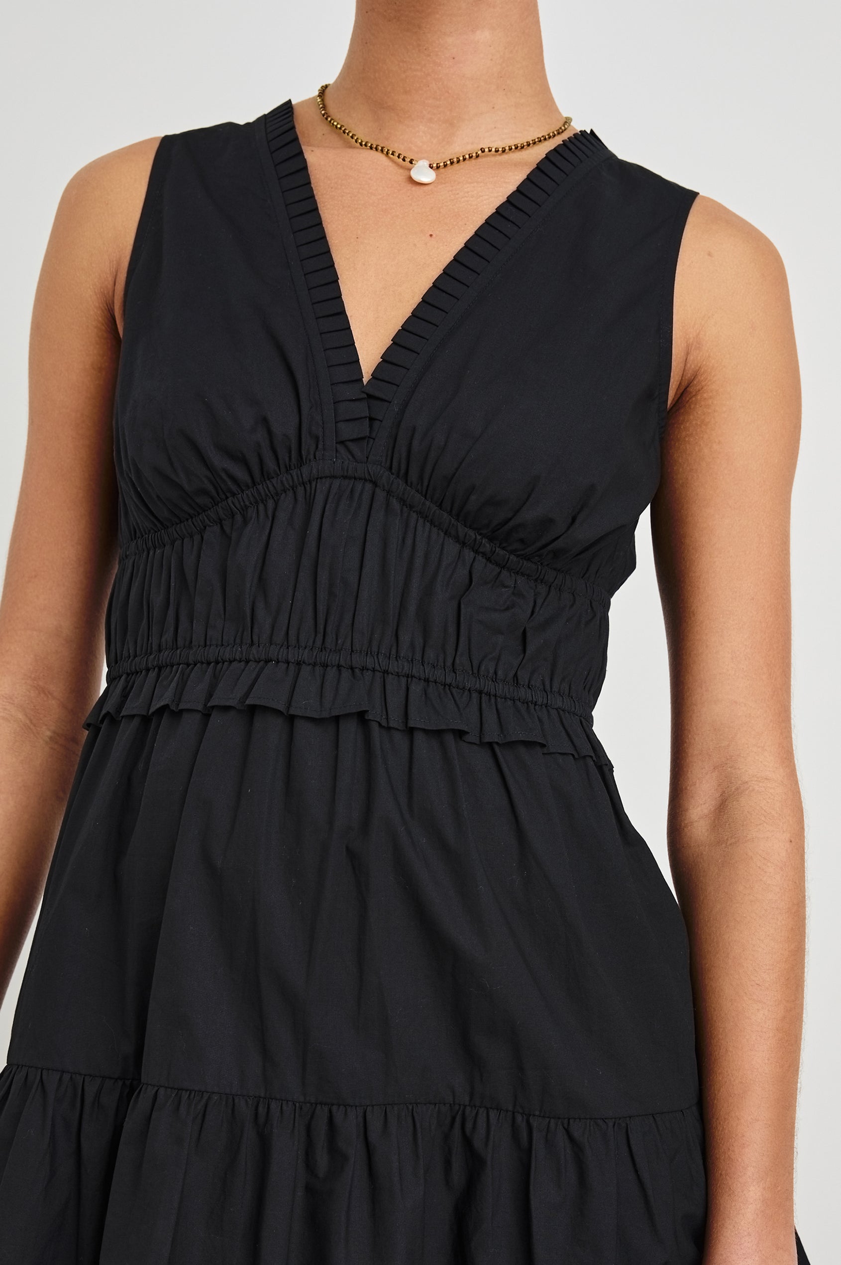 Zoomed in, detail view of female model wearing our sleeveless, Talulah Mini Dress in color Black. Dress features pleated v-neck and smocked waist.