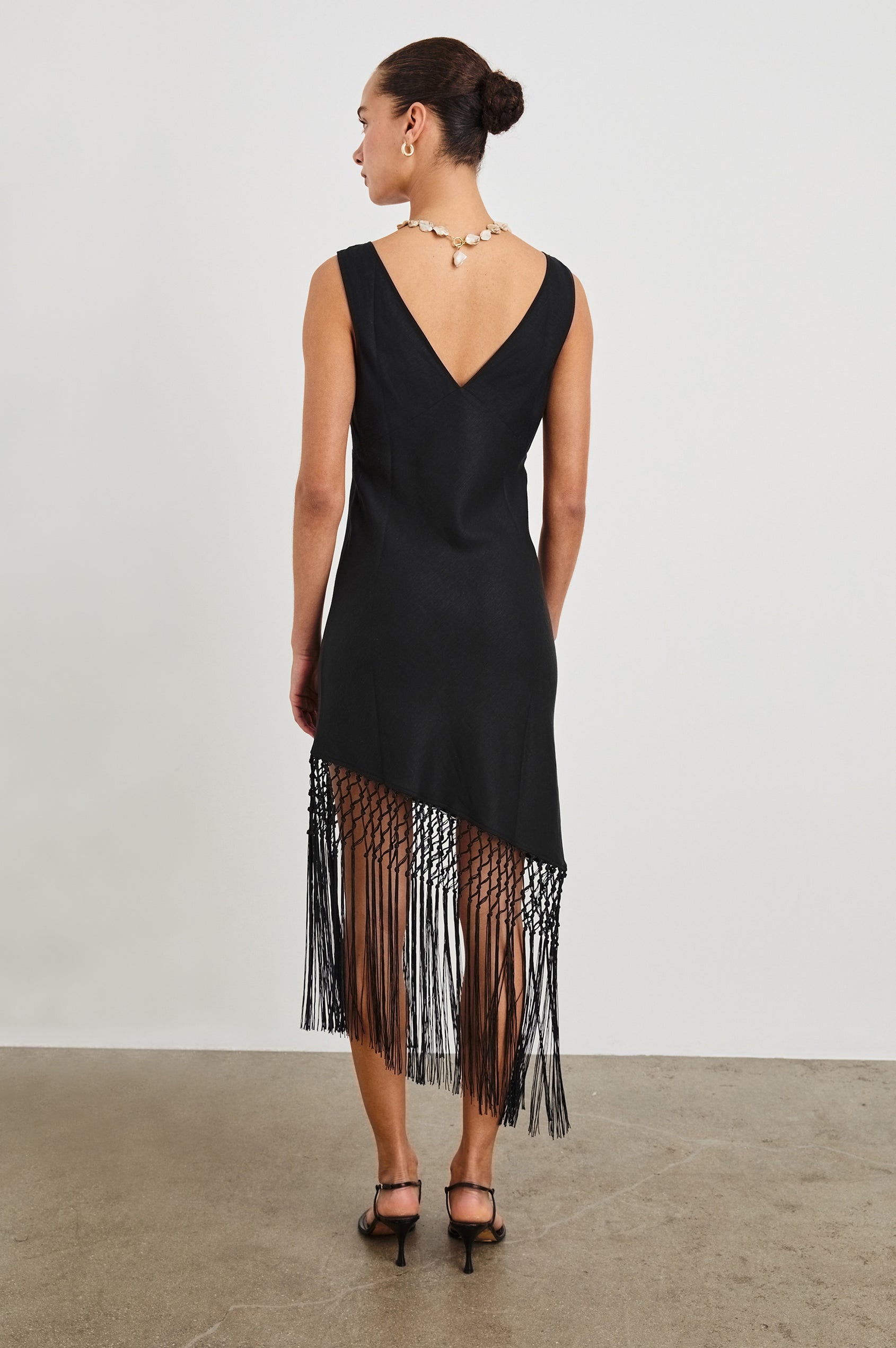 Back view of female model wearing our Tamara Dress in Black.  This is a black mini dress with an asymmetrical fringe hem, sheer netted panel, and empire waistline. 