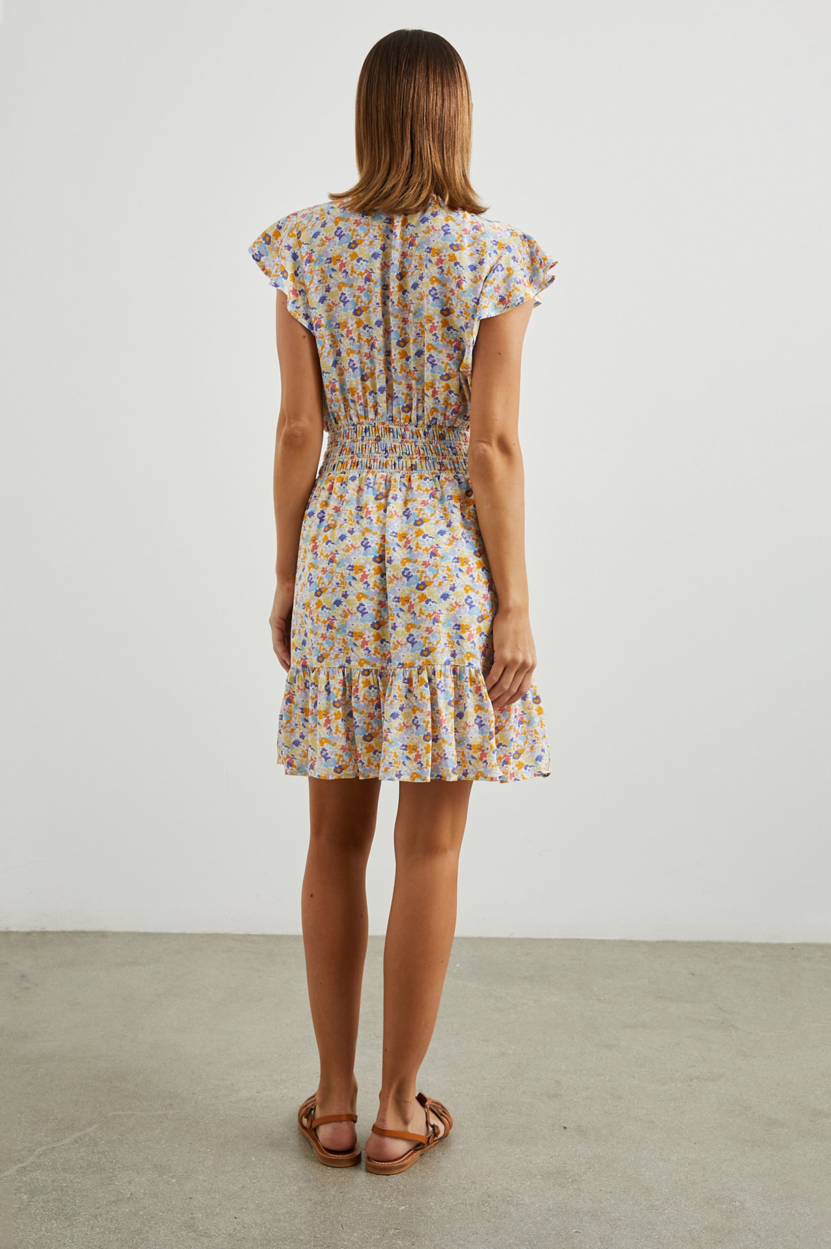 Tara Short Sleeve, Above the Knee Dress with a Smocked Waist, V Neck, Flutter Cap Sleeves, and Tiered Skirt with a Ruffled Hem in Papaya Flowers Colorway, in Orange, Purple, Yellow and Blue - Back Full Body View Featured on Model 