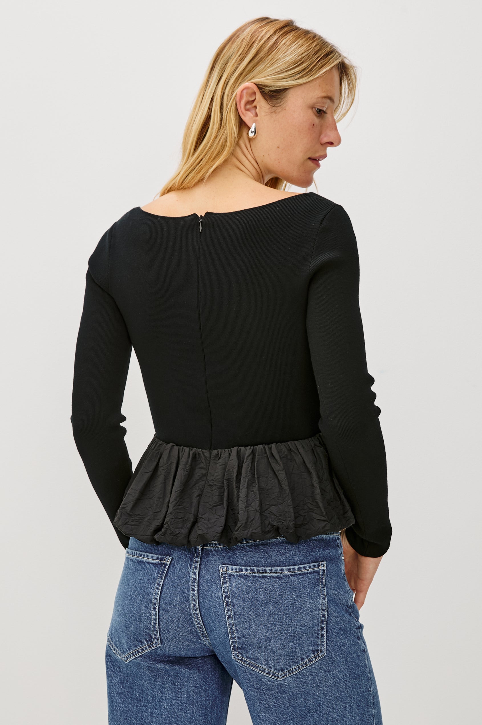 The Tillie is a fitted, long-sleeved sweater top with a contrasting cotton poplin ruffle at the hem in Black Colorway - Back View featured on Model
