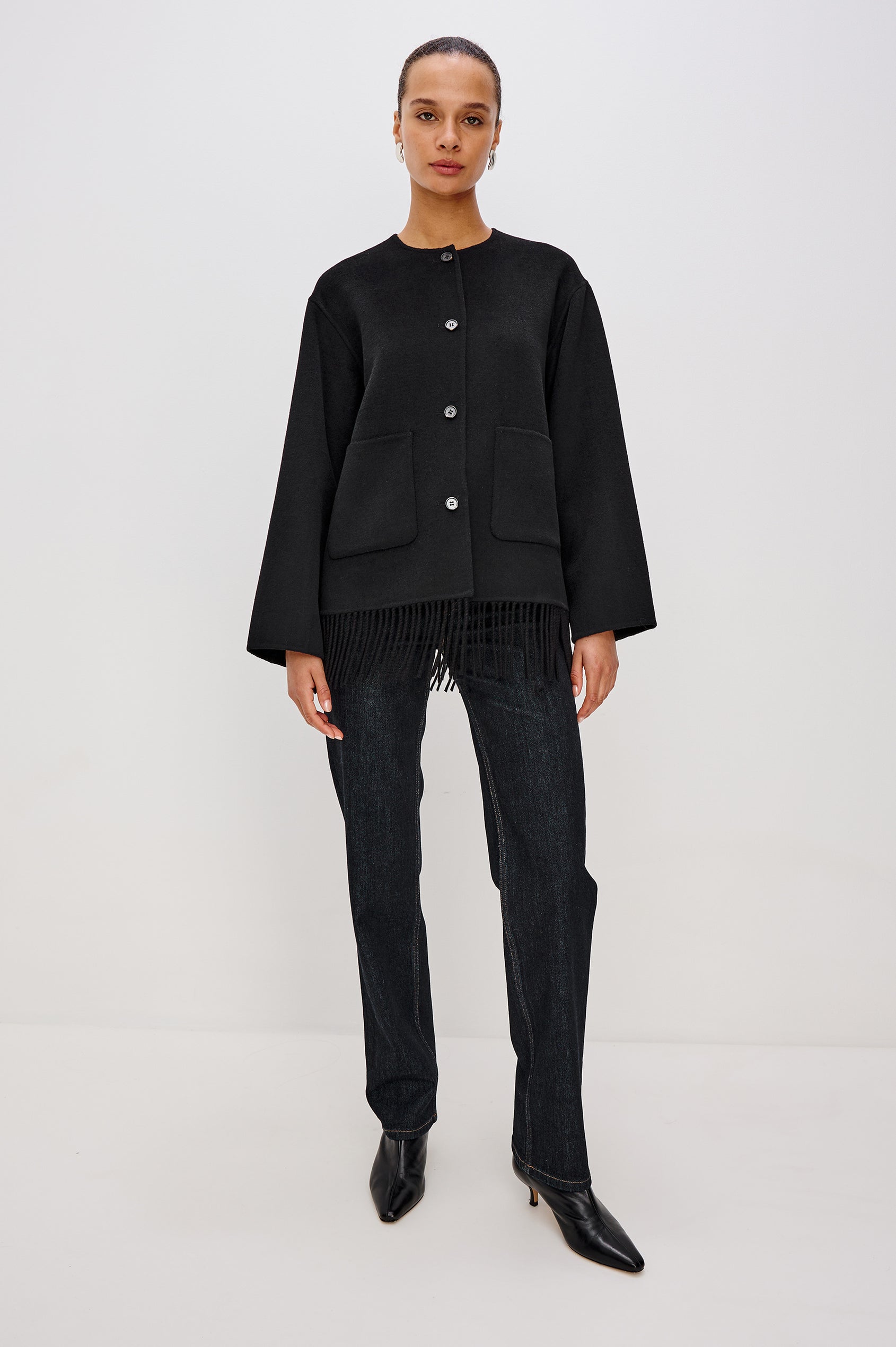 A heavyweight black wool-blend jacket with a relaxed fit. The jacket includes front button closures, patch pockets, and fringe detail at the hem for added texture and style. Full Body Front View Featured on Model.