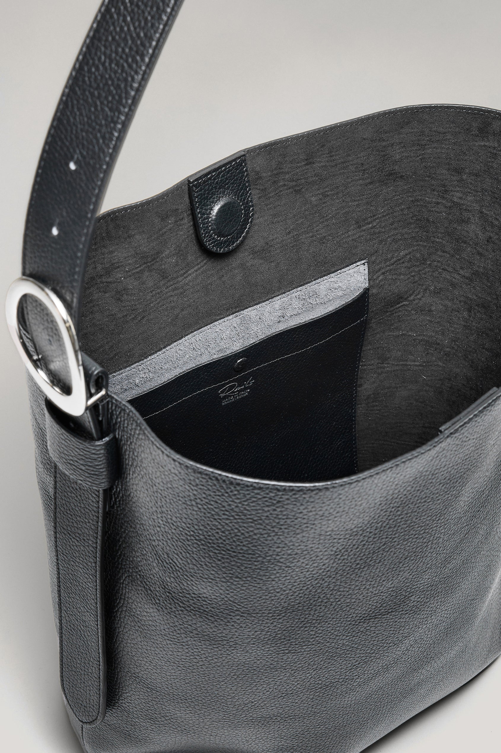 The Torino is a soft pebbled leather bucket bag, designed with an adjustable strap and polished silver oval buckle, in black colorway. Top view of interior featured.