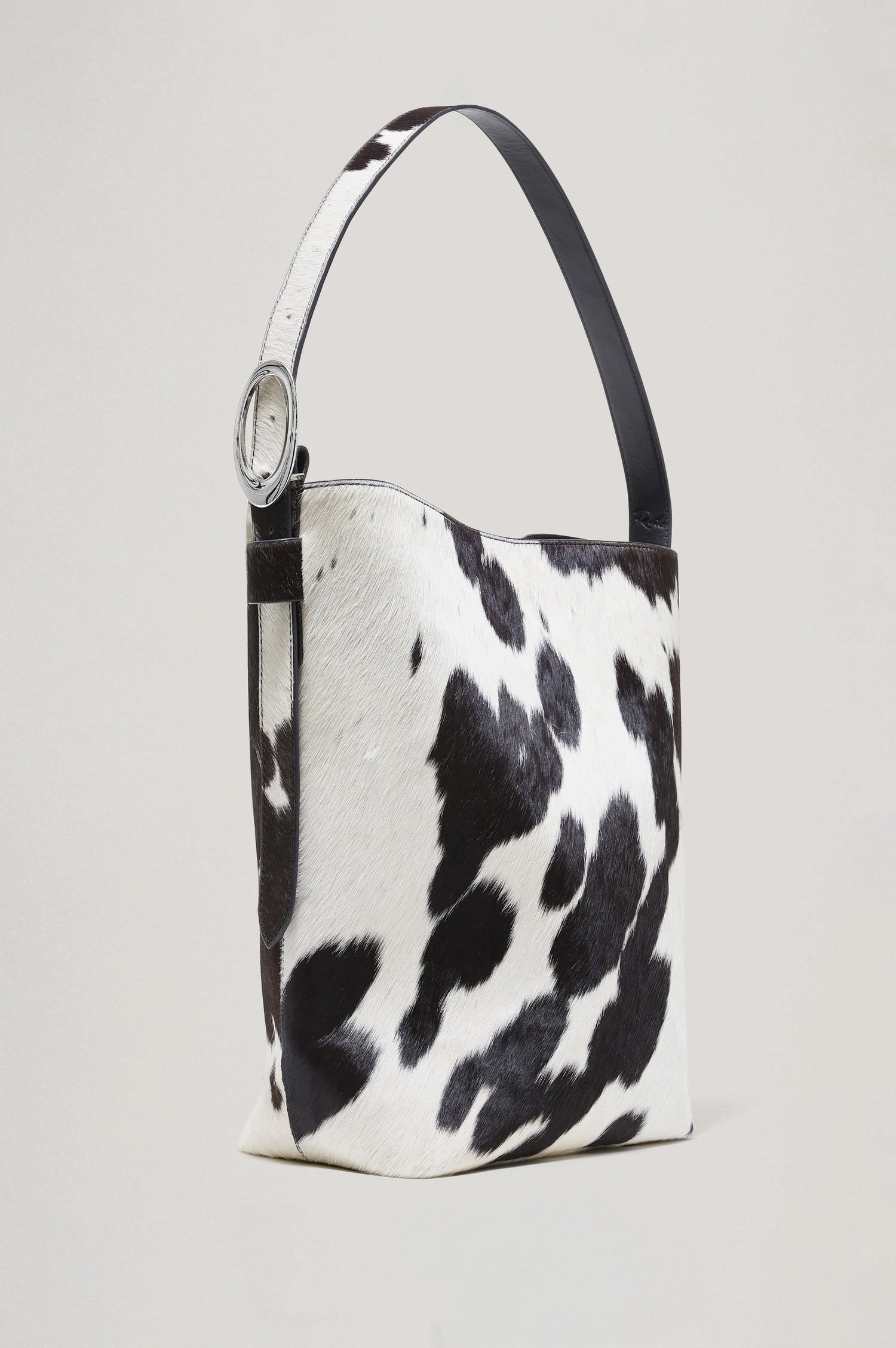 The Torino is a cow-print haircalf leather bucket bag, designed with an adjustable strap and polished silver oval buckle. Front view featured.