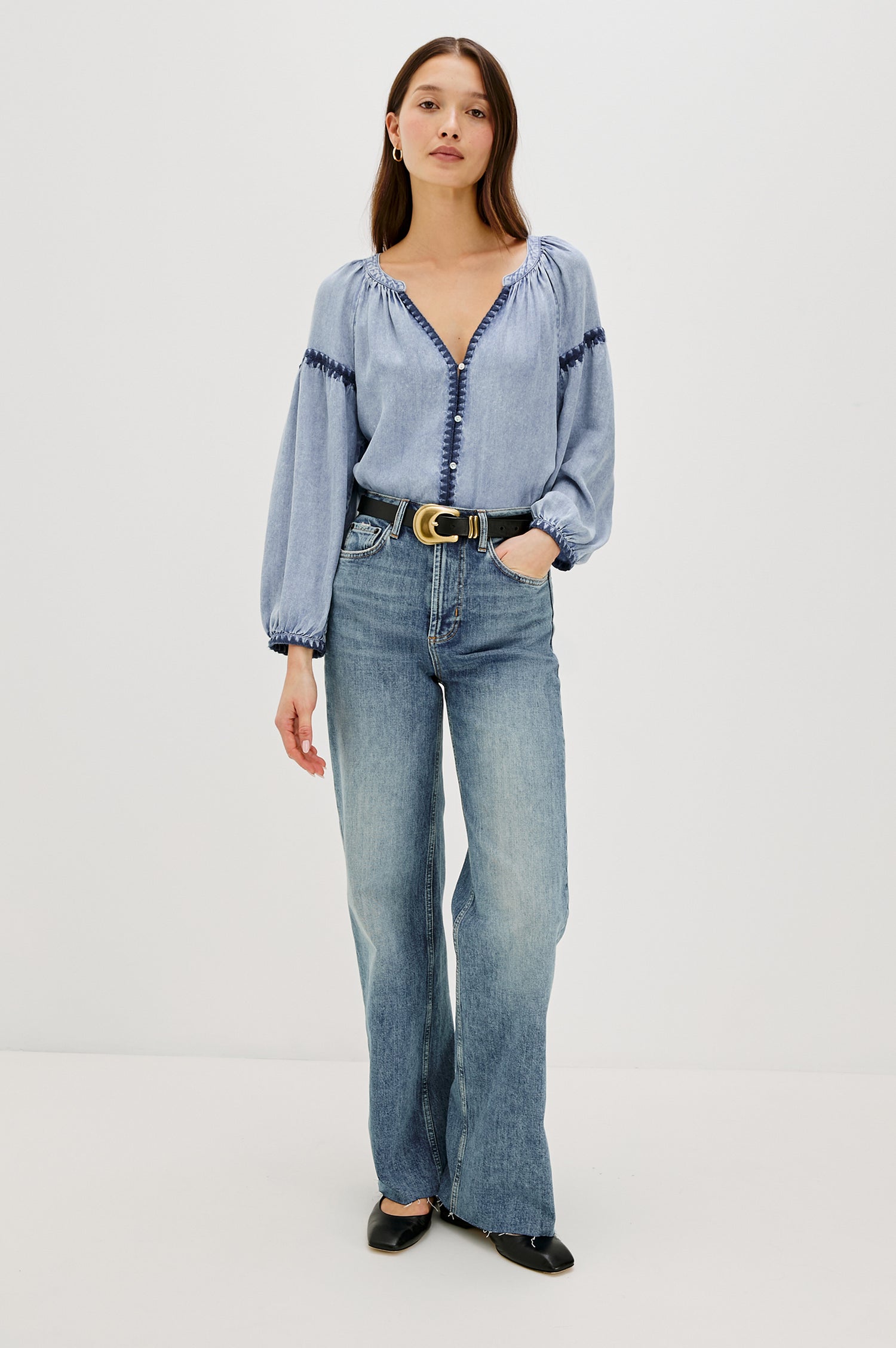 Toshi loose-fitting long sleeve top featuring a v-neckline, front button closure in Medium Cloud Wash Colorway. Contrast blanket stitching at the front, below shoulder sleeve and at the cuff - full body front view on model, shirt tucked in with belt