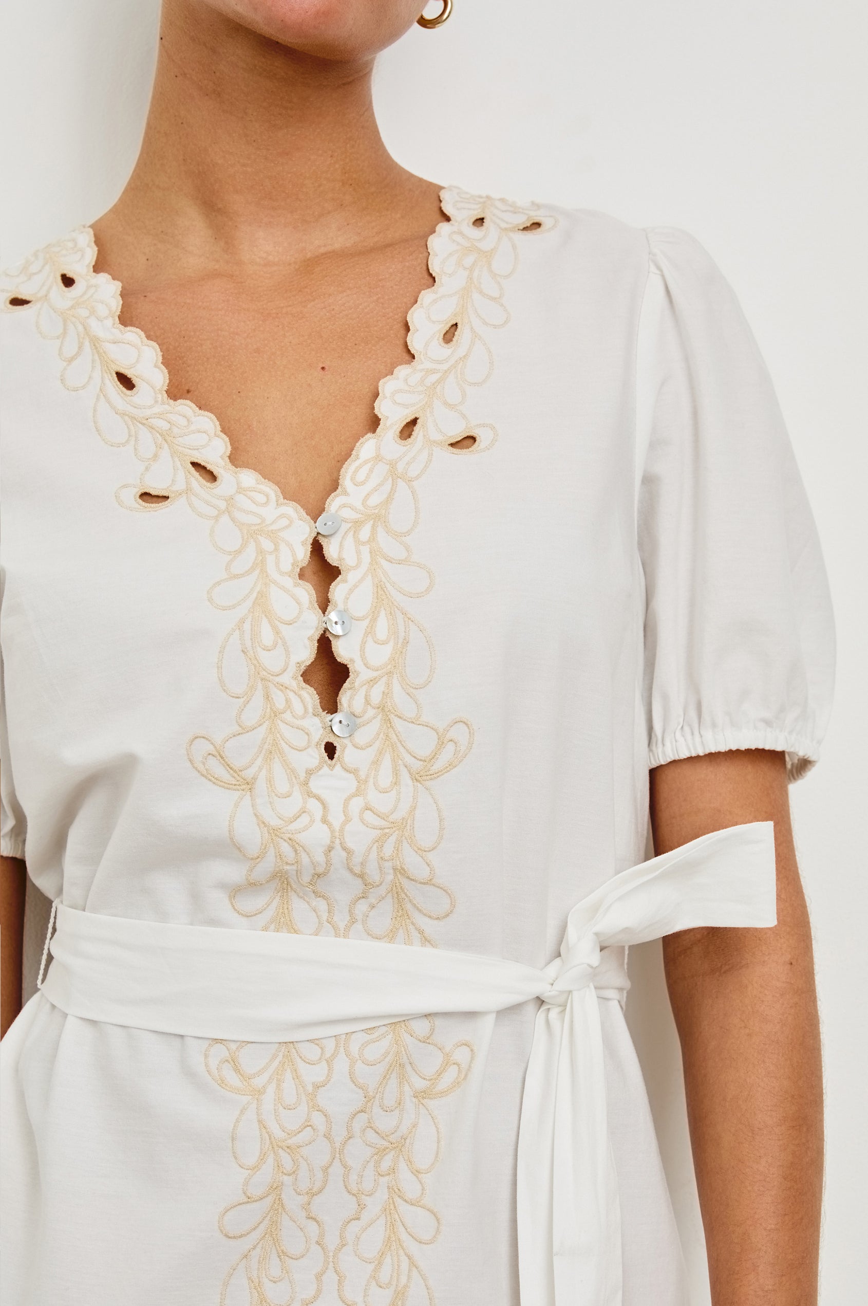 Detail view of model wearing a white mini summer dress with peplum sleeves and contrast eyelet embroider. Dress has a belt that can be tied at the front or back. Features 3 buttons in the front.