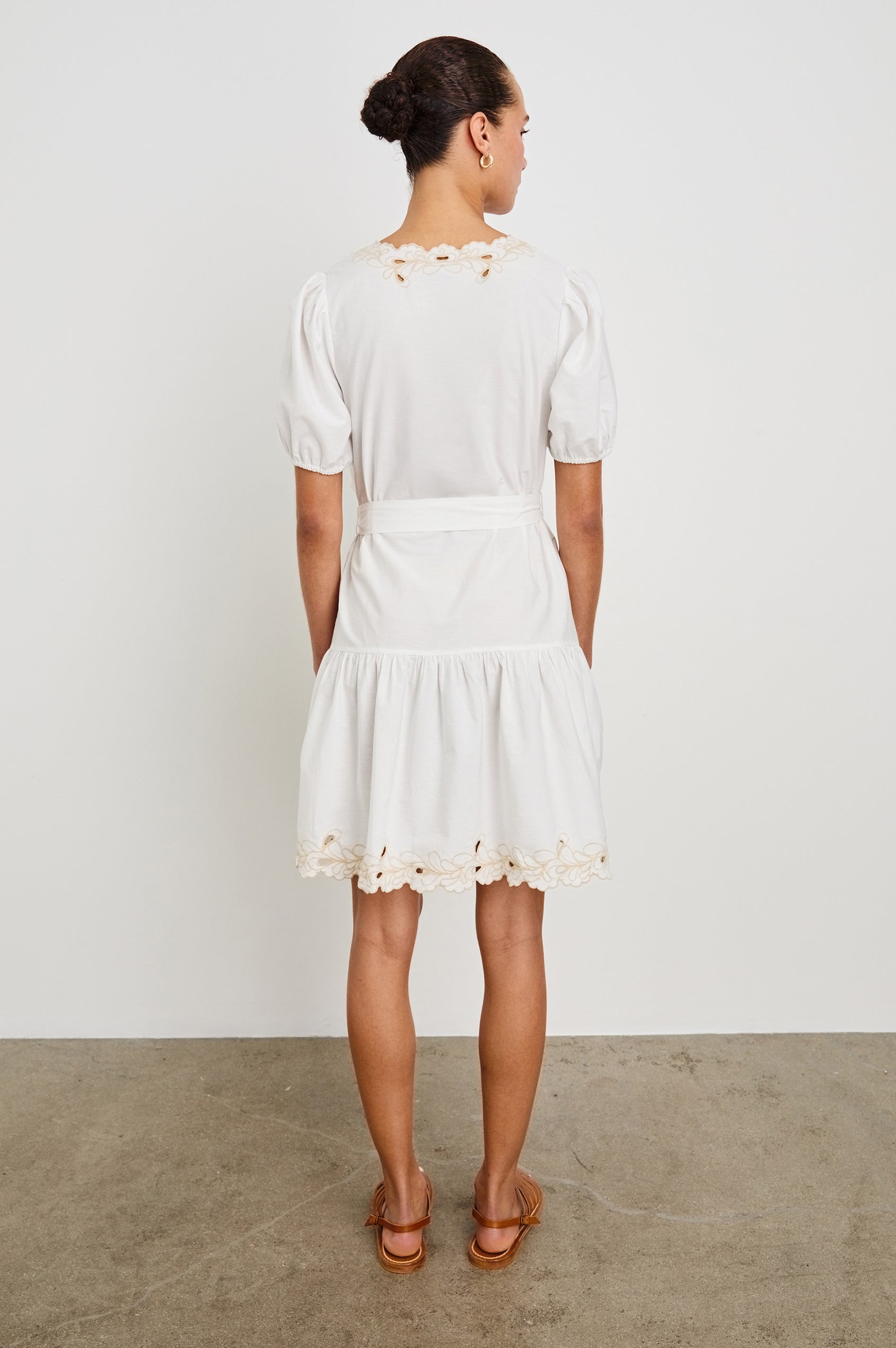 Back view of model wearing a white mini summer dress with peplum sleeves and contrast eyelet embroider. Dress has a belt that can be tied at the front or back. Styled with tan colored sandals. 