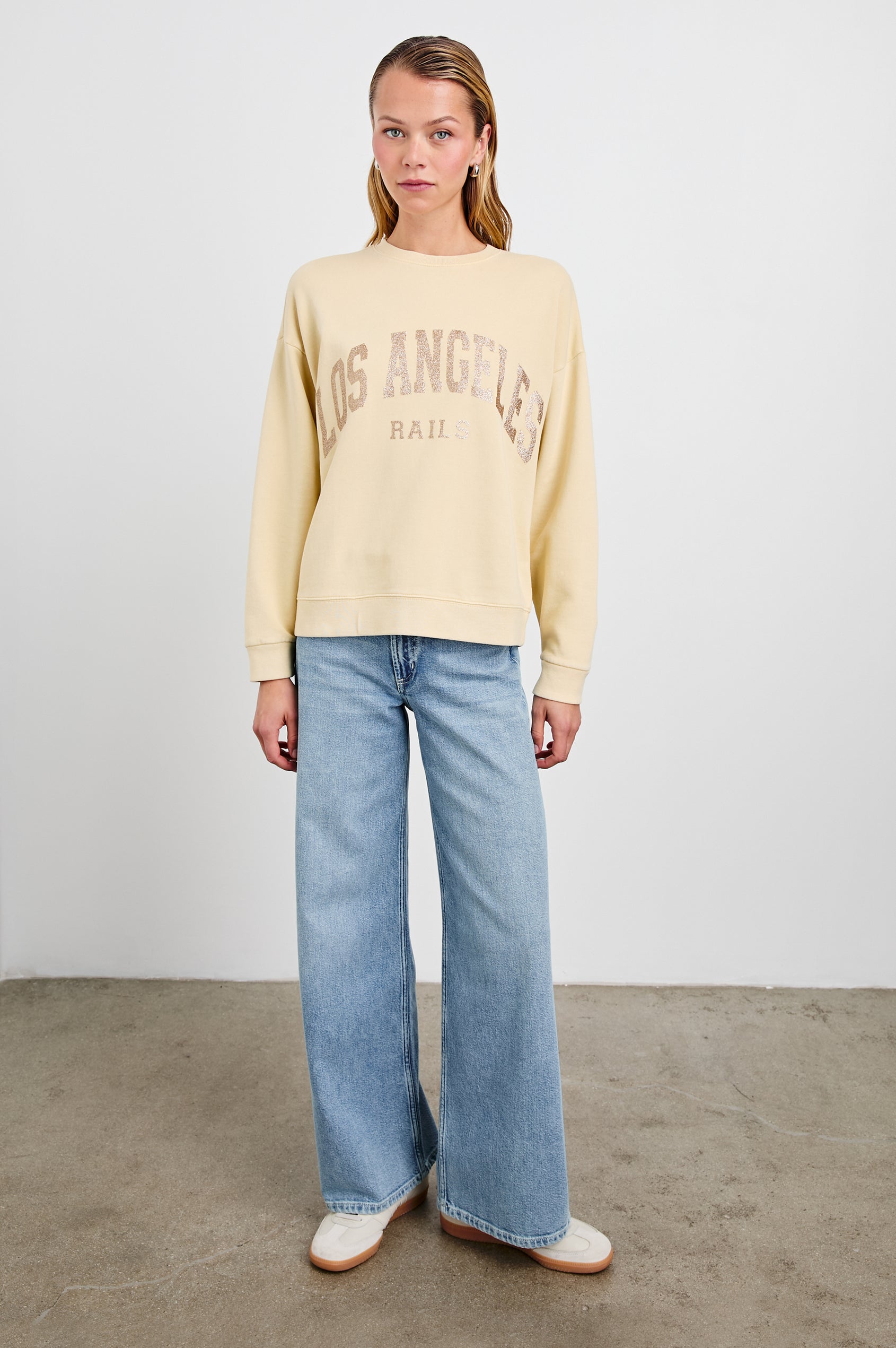 The Varsity Sweatshirt with a Crew Neck and Gold Glitter Text states Los Angeles Rails in Los Angeles Gold Glitter Colorway, on pale yellow - Front Full Body View Featured on Model 