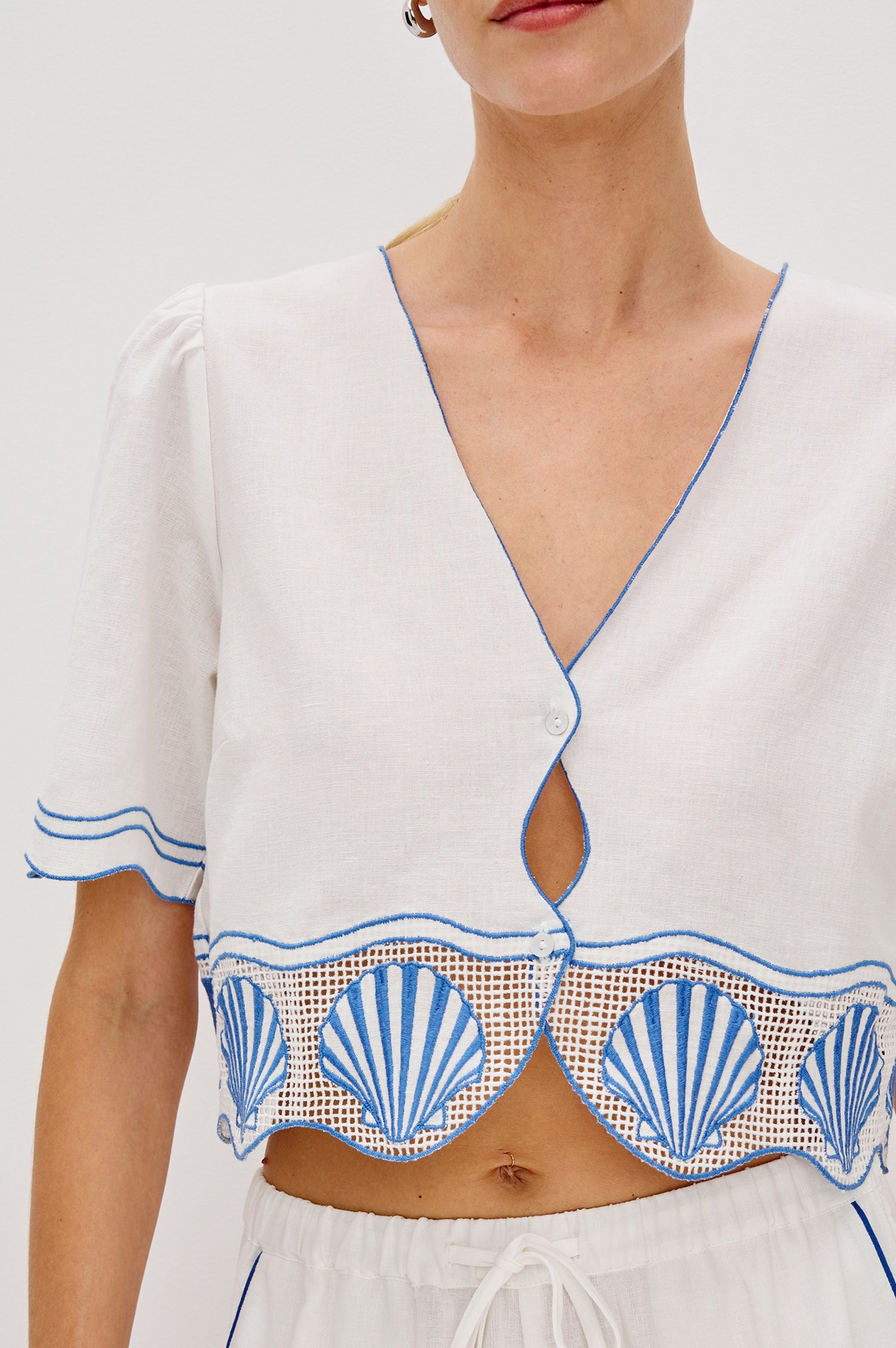 Venice Cropped V-Neck Top with Short Sleeves (slightly puffed), 2-Button Front, Keyhole at Center Front, Contrast Blue Sea Shell Embroidery and Scalloped Hem in White Colorway. Front Detail View Shown on Model.