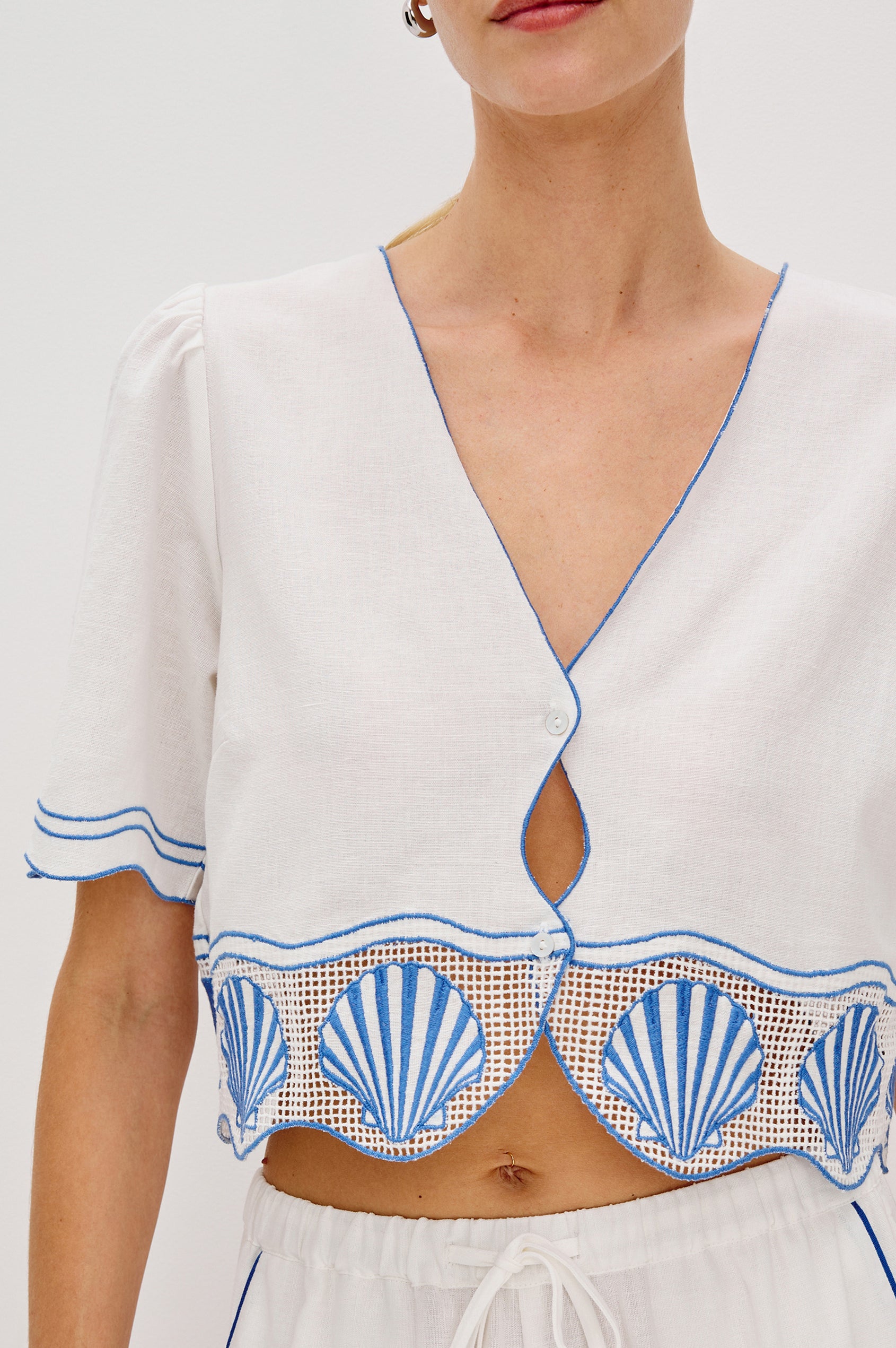 Venice Cropped V-Neck Top with Short Sleeves (slightly puffed), 2-Button Front, Keyhole at Center Front, Contrast Blue Sea Shell Embroidery and Scalloped Hem in White Colorway. Front Detail View Shown on Model.