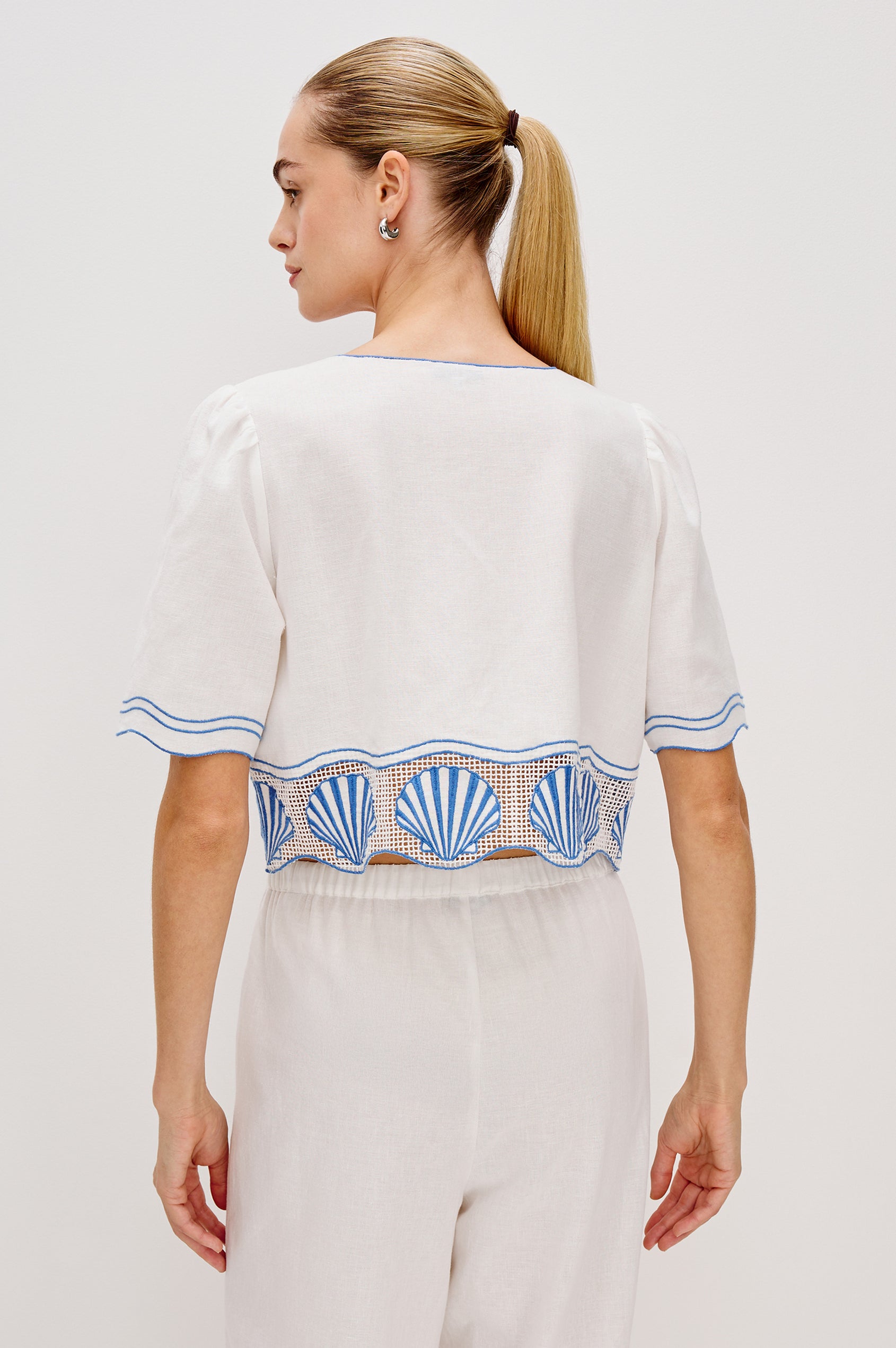 Venice Cropped V-Neck Top with Short Sleeves (slightly puffed), 2-Button Front, Keyhole at Center Front, Contrast Blue Sea Shell Embroidery and Scalloped Hem in White Colorway. Back View Shown on Model.
