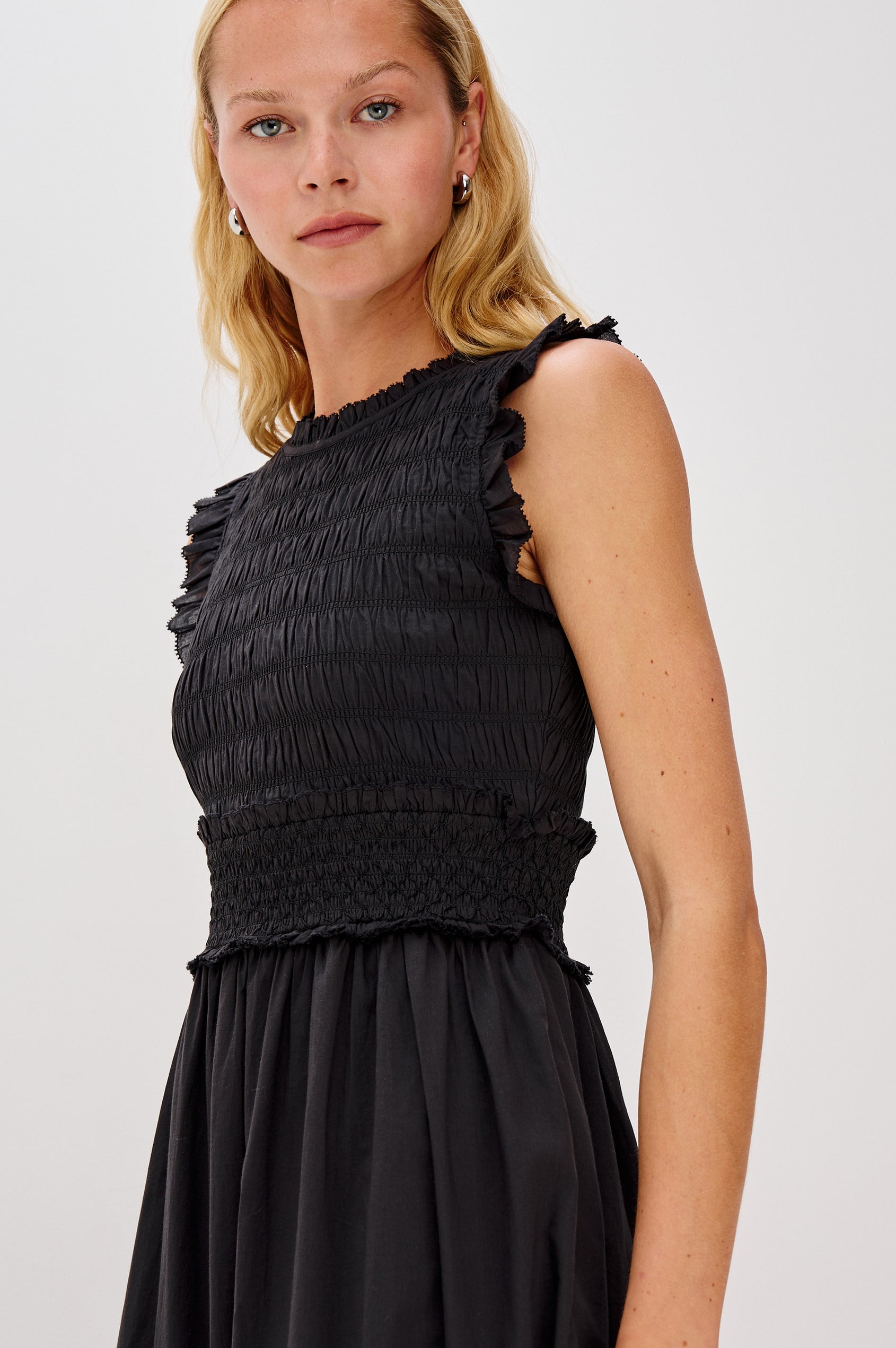 Vivier is a crinkled cotton poplin maxi dress featuring a smocked, fitted bodice and waistband. The dress flows into a relaxed skirt with soft ruffled tiers, creating a breezy silhouette in Black Colorway - Close up of upper Details on Model