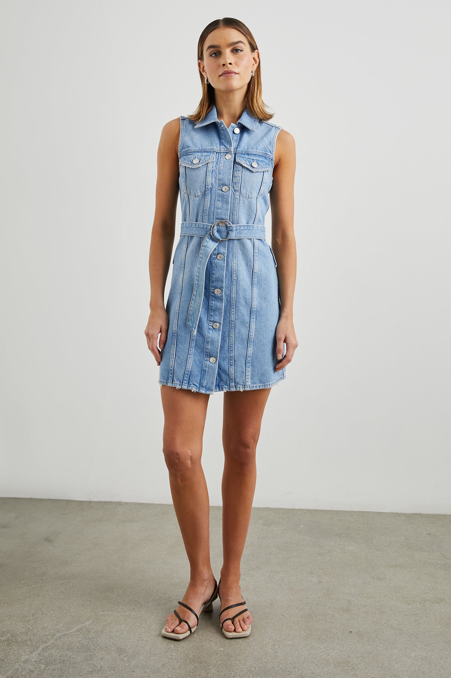 Wilshire Sleeveless Denim Mini Dress with a Full Button Front, Two Chest Button Flap Pockets, a Double Ring Belt, a Collar, and Front Vertical Seams in Blue Denim Colorway - Front Full Body View Featured on Model 