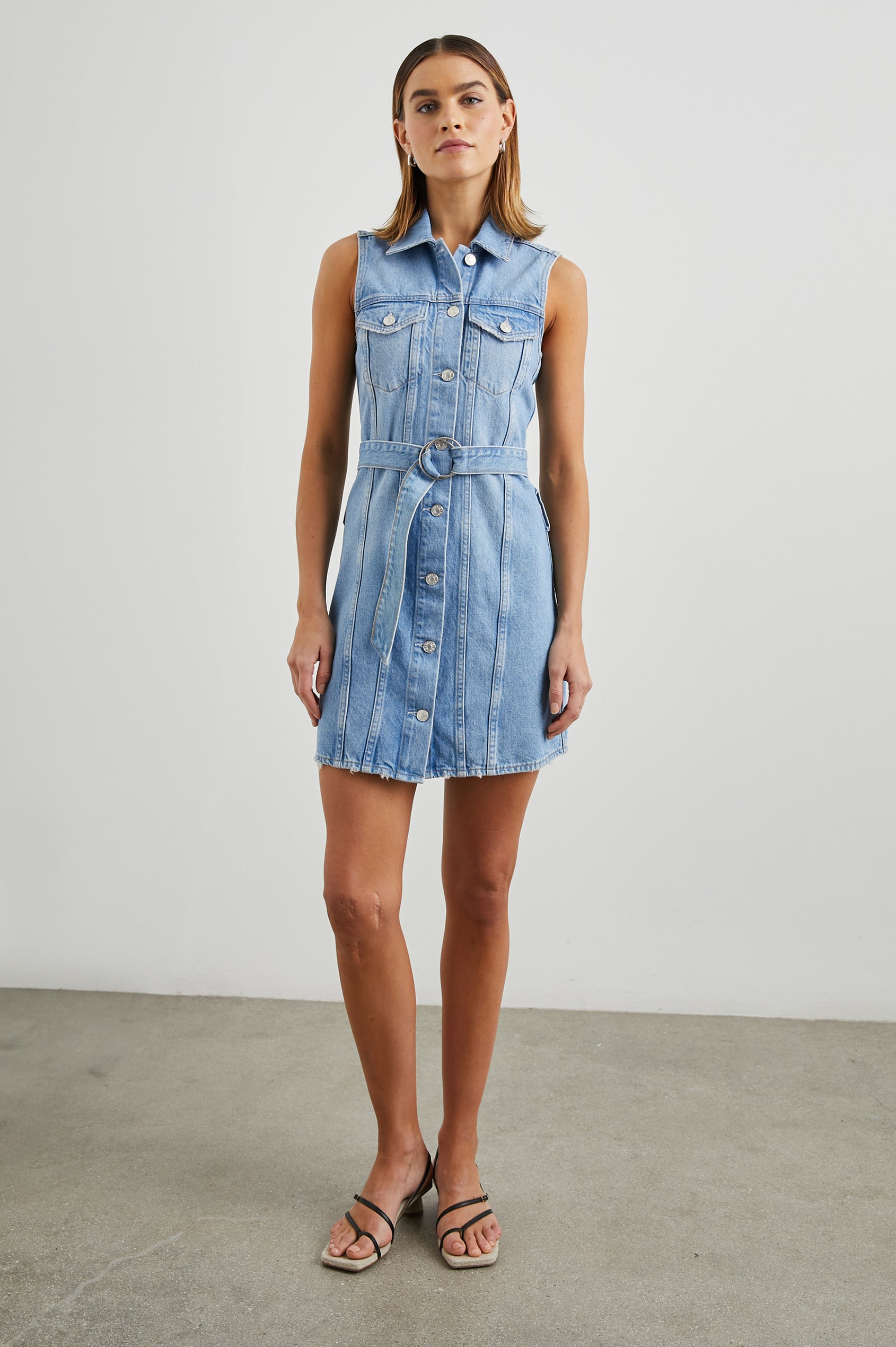 Wilshire Sleeveless Denim Mini Dress with a Full Button Front, Two Chest Button Flap Pockets, a Double Ring Belt, a Collar, and Front Vertical Seams in Blue Denim Colorway - Front Full Body View Featured on Model 
