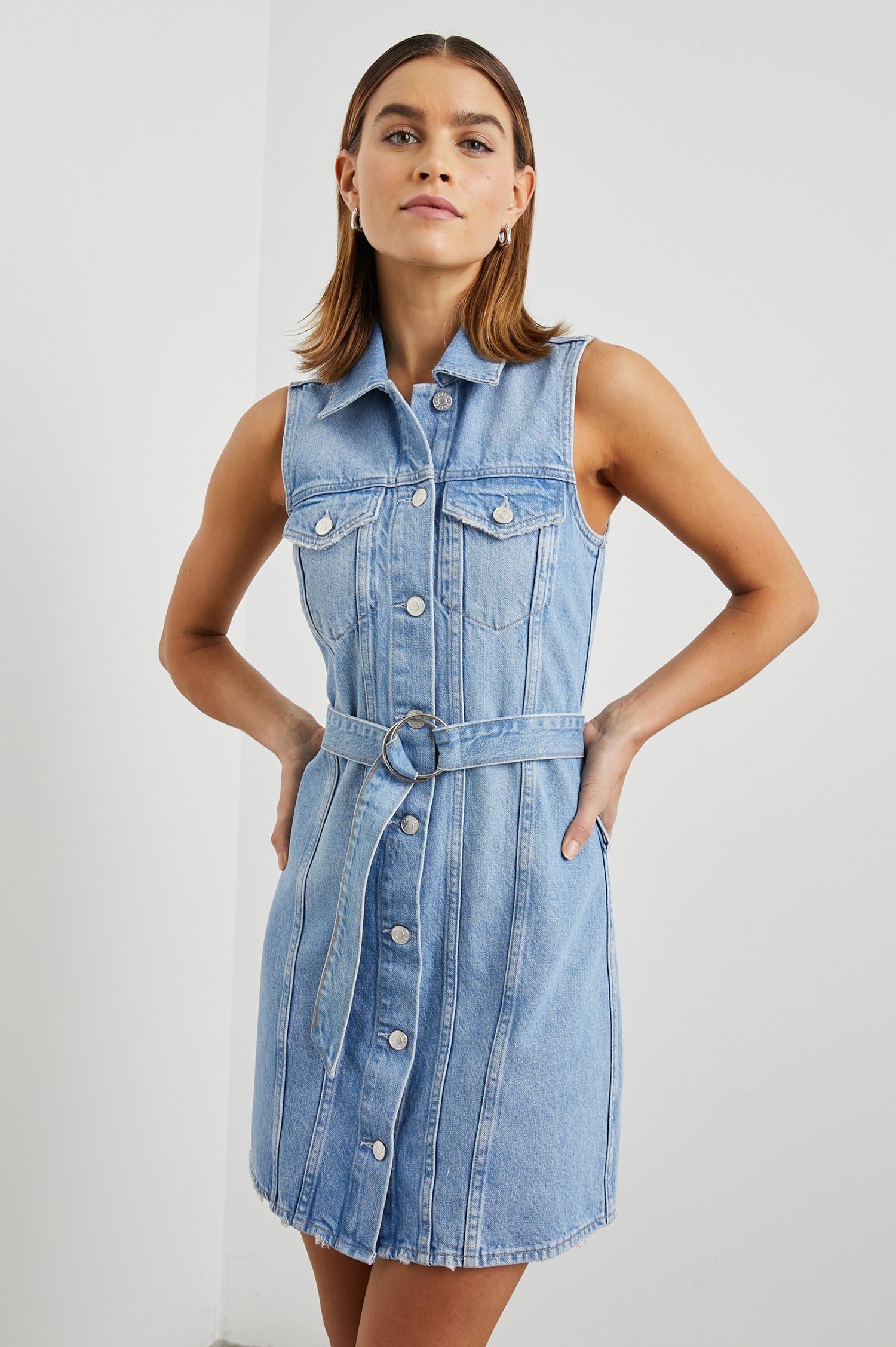 Wilshire Sleeveless Denim Mini Dress with a Full Button Front, Two Chest Button Flap Pockets, a Double Ring Belt, a Collar, and Front Vertical Seams in Blue Denim Colorway - Front View Featured on Model 