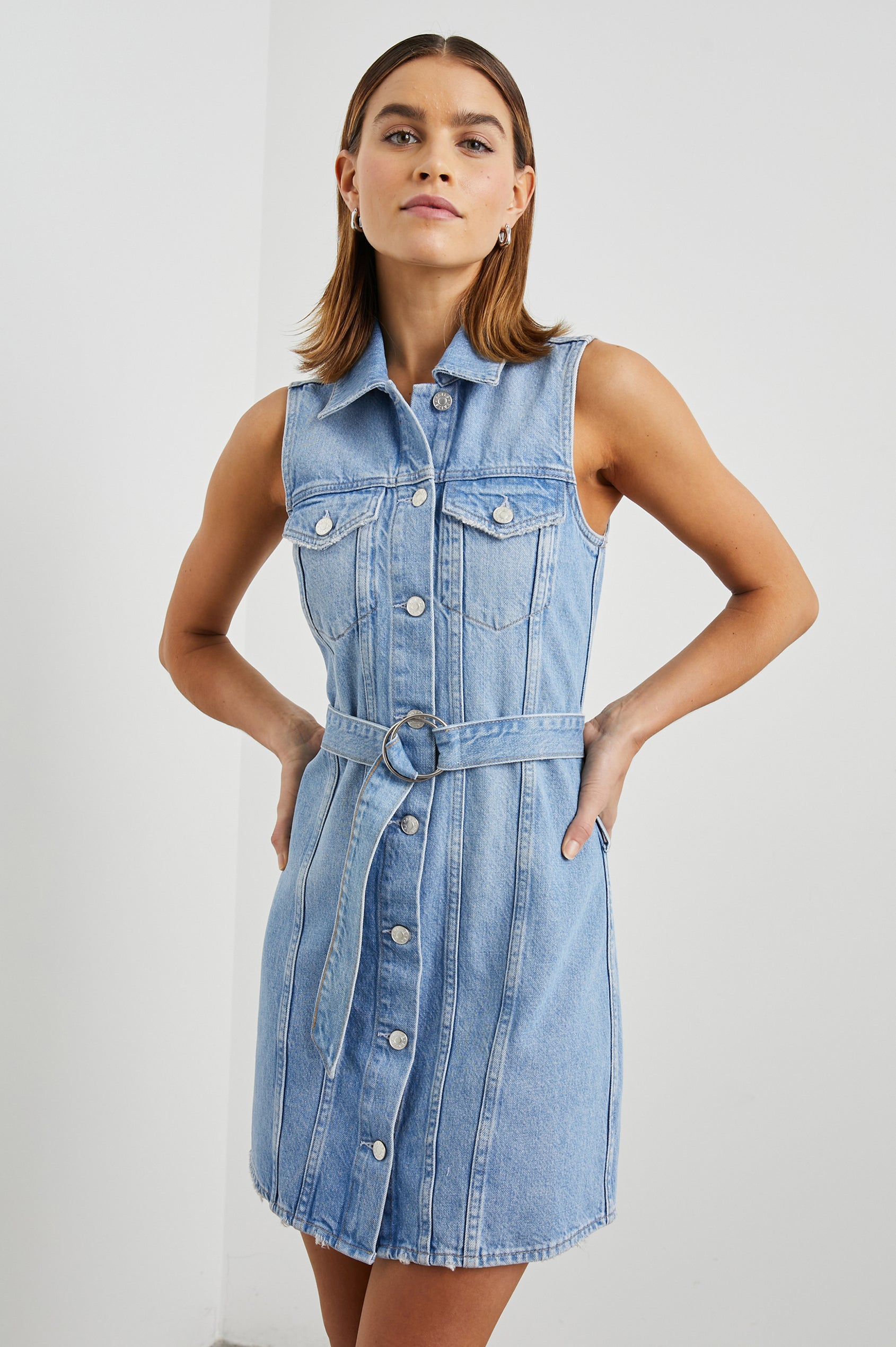 Wilshire Sleeveless Denim Mini Dress with a Full Button Front, Two Chest Button Flap Pockets, a Double Ring Belt, a Collar, and Front Vertical Seams in Blue Denim Colorway - Front View Featured on Model 