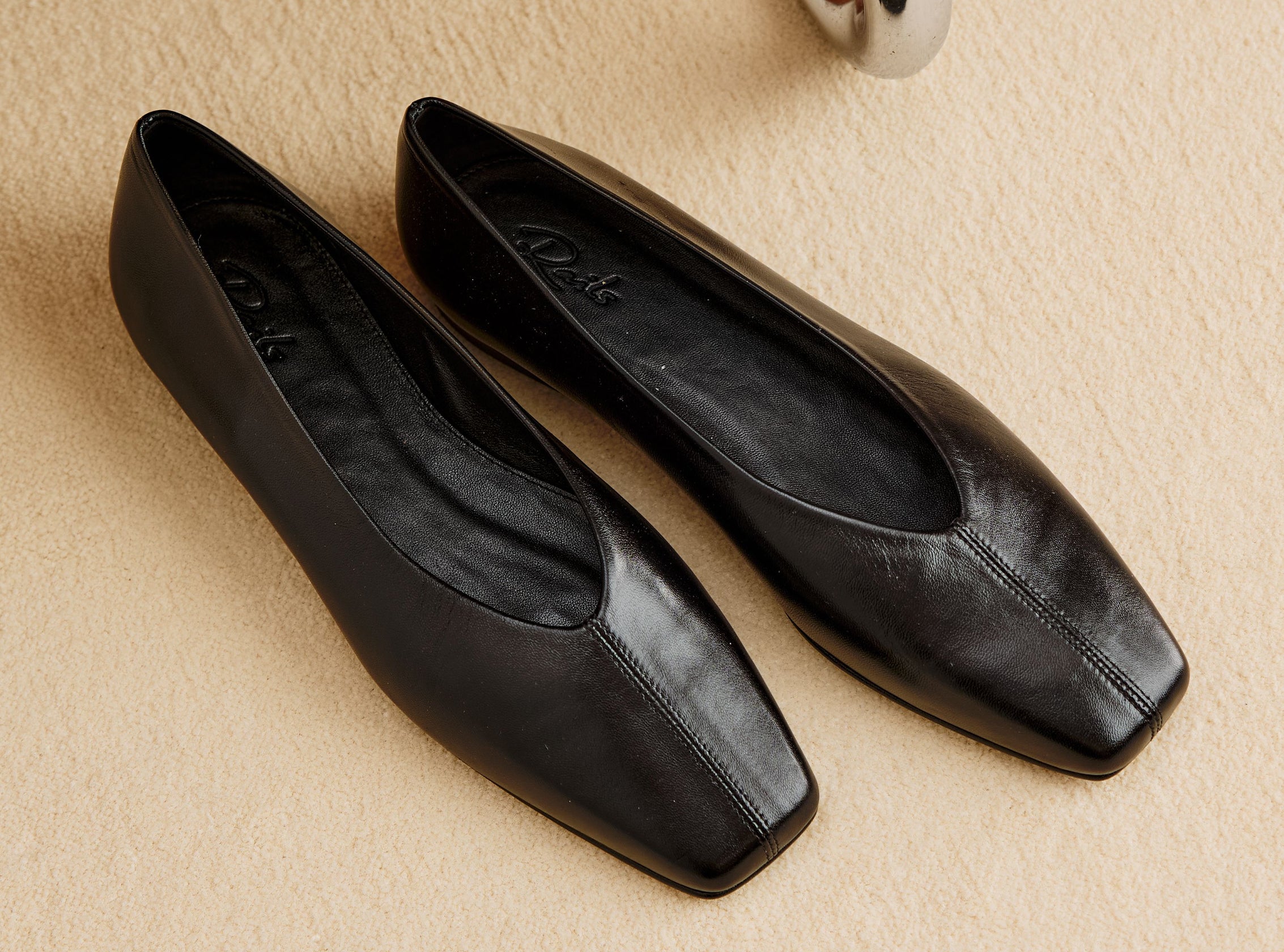 A pair of black leather square-toe flats with a low block heel, displayed on a textured beige surface.