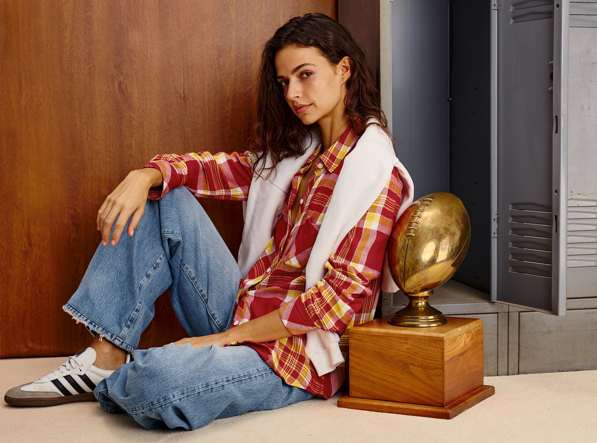 A woman sits on a floor in a locker-room-like setting, wearing a red and yellow plaid flannel shirt, blue jeans, a white zip-front vest, and white sneakers. A gold football trophy on a wooden base sits beside her.