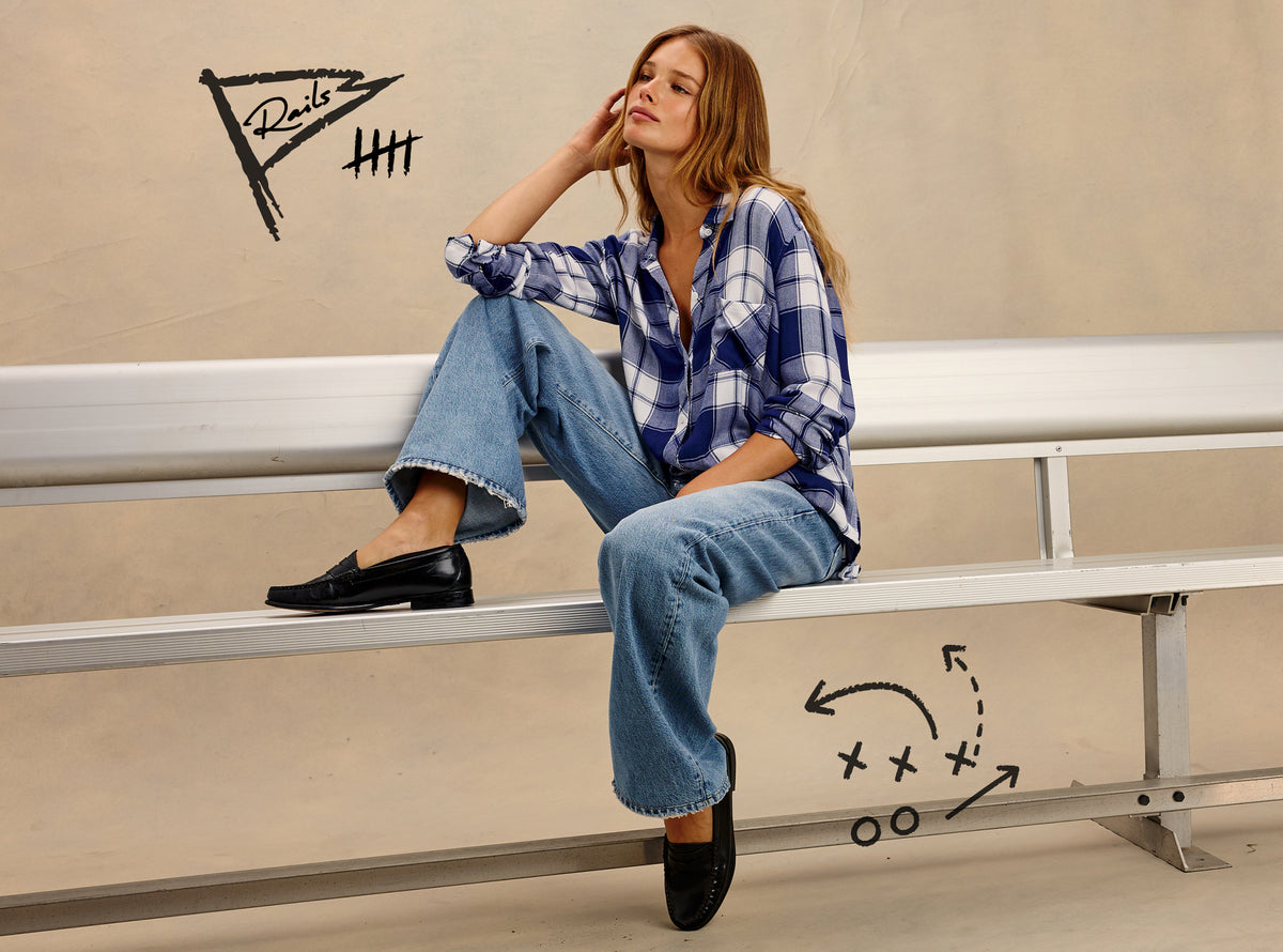 A woman sits on metal stadium seating wearing a blue and white plaid button-up shirt, wide-leg blue jeans, and black loafers. Hand-drawn sports-style graphics and the word 'Rails' appear on the beige wall behind her.