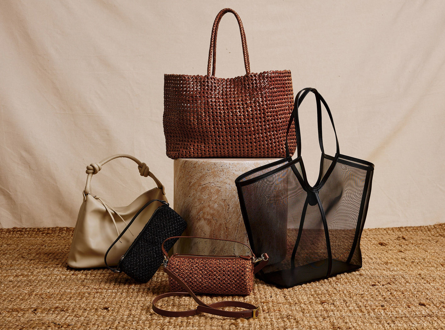 A styled product display of five handbags on a woven jute surface against a draped beige fabric backdrop. 