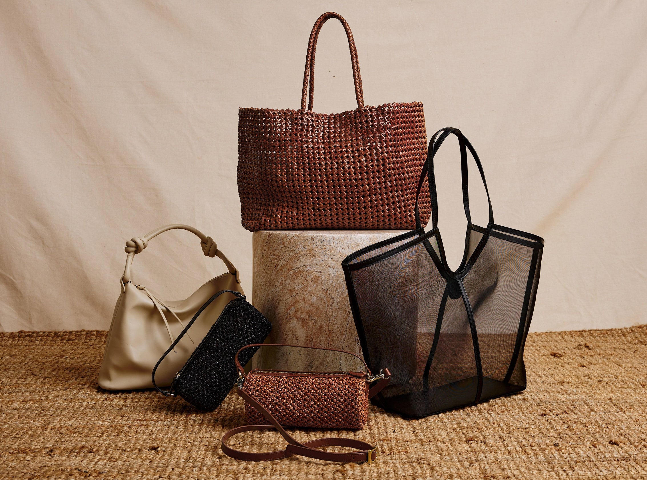 A styled product display of five handbags on a woven jute surface against a draped beige fabric backdrop. 