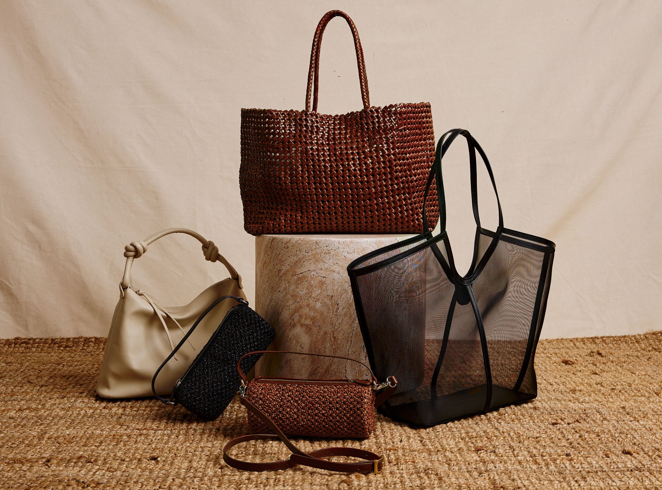 Collection of handbags including a woven brown bag, a black mesh tote, a beige bucket bag, and a small brown purse on a textured surface.