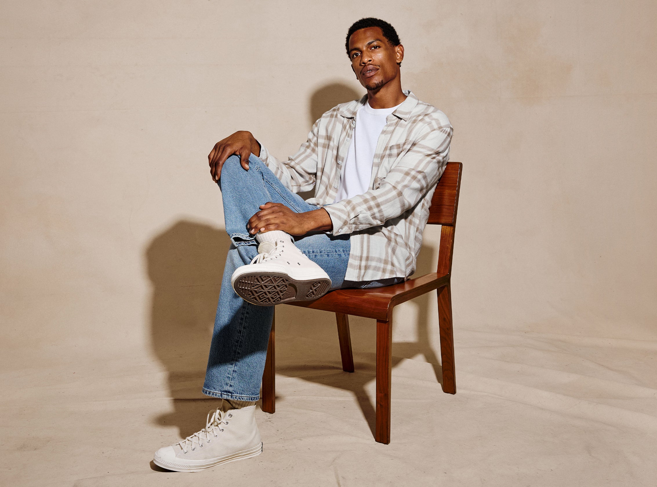 A man sits on a wooden chair wearing a beige and white plaid button-up shirt over a white t-shirt, blue jeans, and white high-top sneakers against a warm beige studio backdrop.