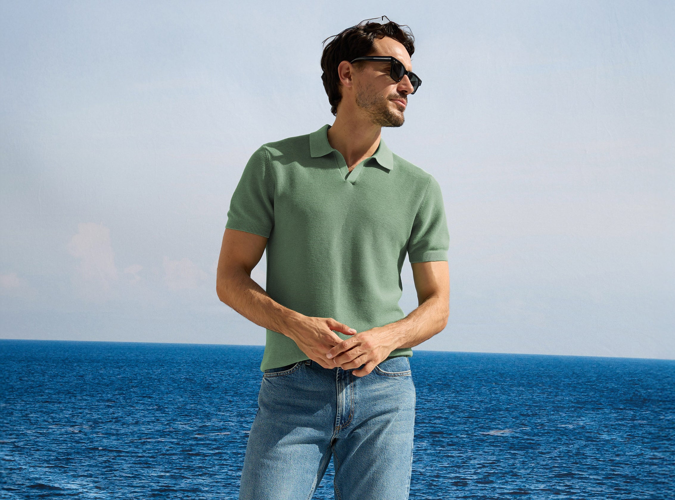 Man wearing a green polo shirt and sunglasses standing by the ocean.