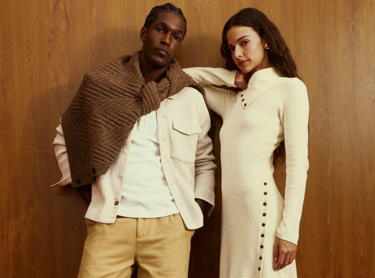 A man and a woman posing together against a warm wood-paneled wall. The man wears a white shirt, beige trousers, and a brown textured knit wrap draped over his shoulders.