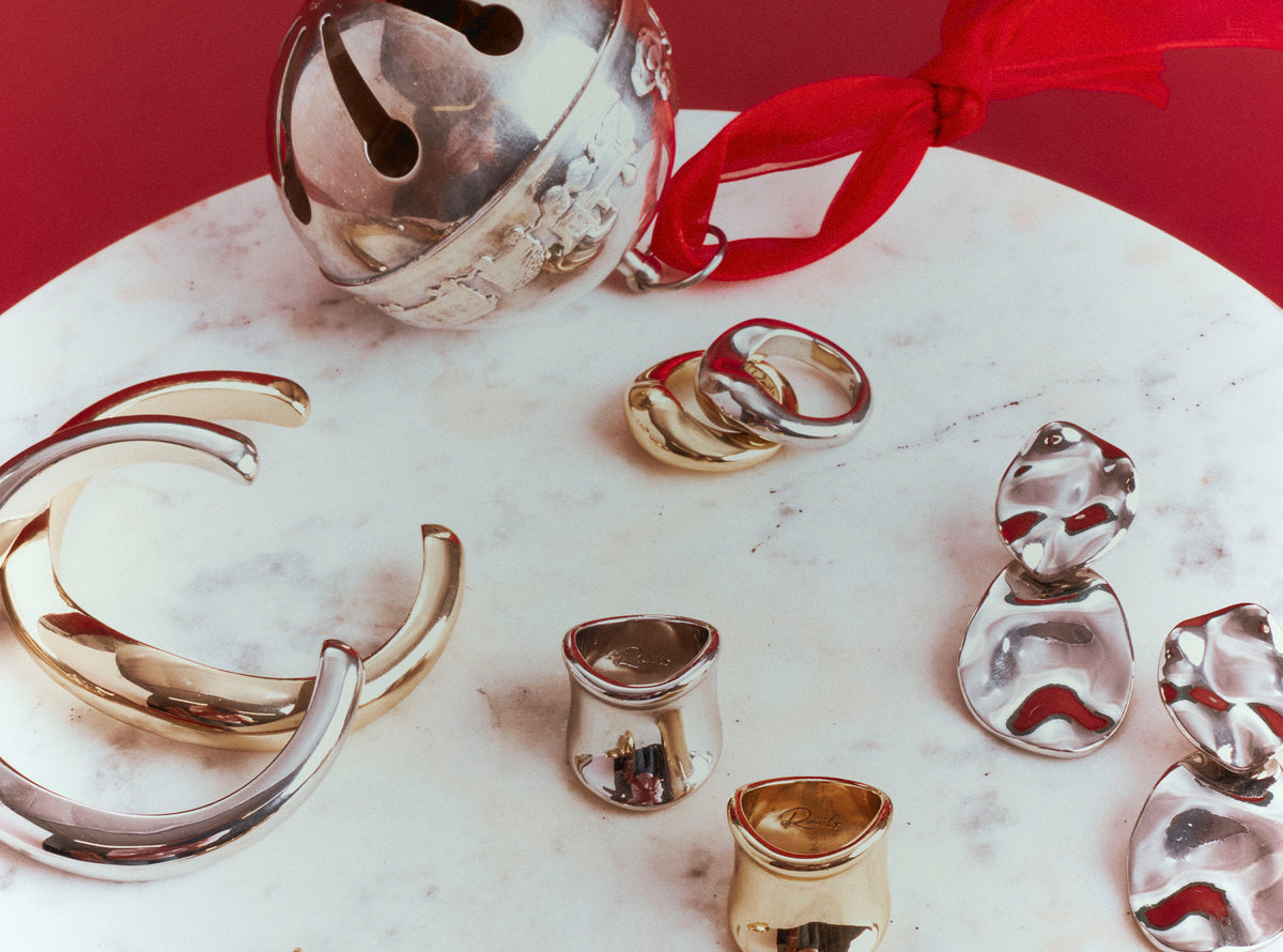 A collection of polished metallic jewelry pieces, including a large silver-toned cuff, a silver bell with a red ribbon, several rings, and small sculptural pieces, arranged on a white marble surface against a solid red background.