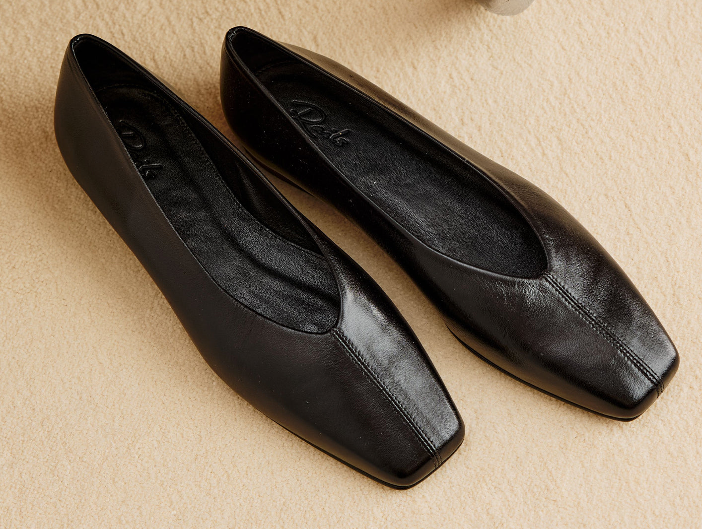 A pair of black leather square-toe flats with a low block heel, displayed on a textured beige surface.