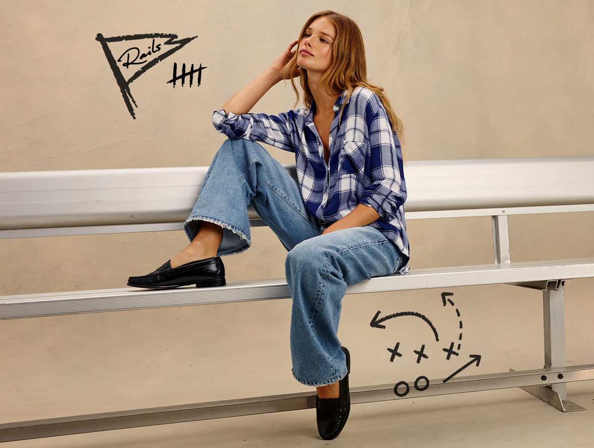 A woman sits on metal stadium seating wearing a blue and white plaid button-up shirt, wide-leg blue jeans, and black loafers. Hand-drawn sports-style graphics and the word 'Rails' appear on the beige wall behind her.
