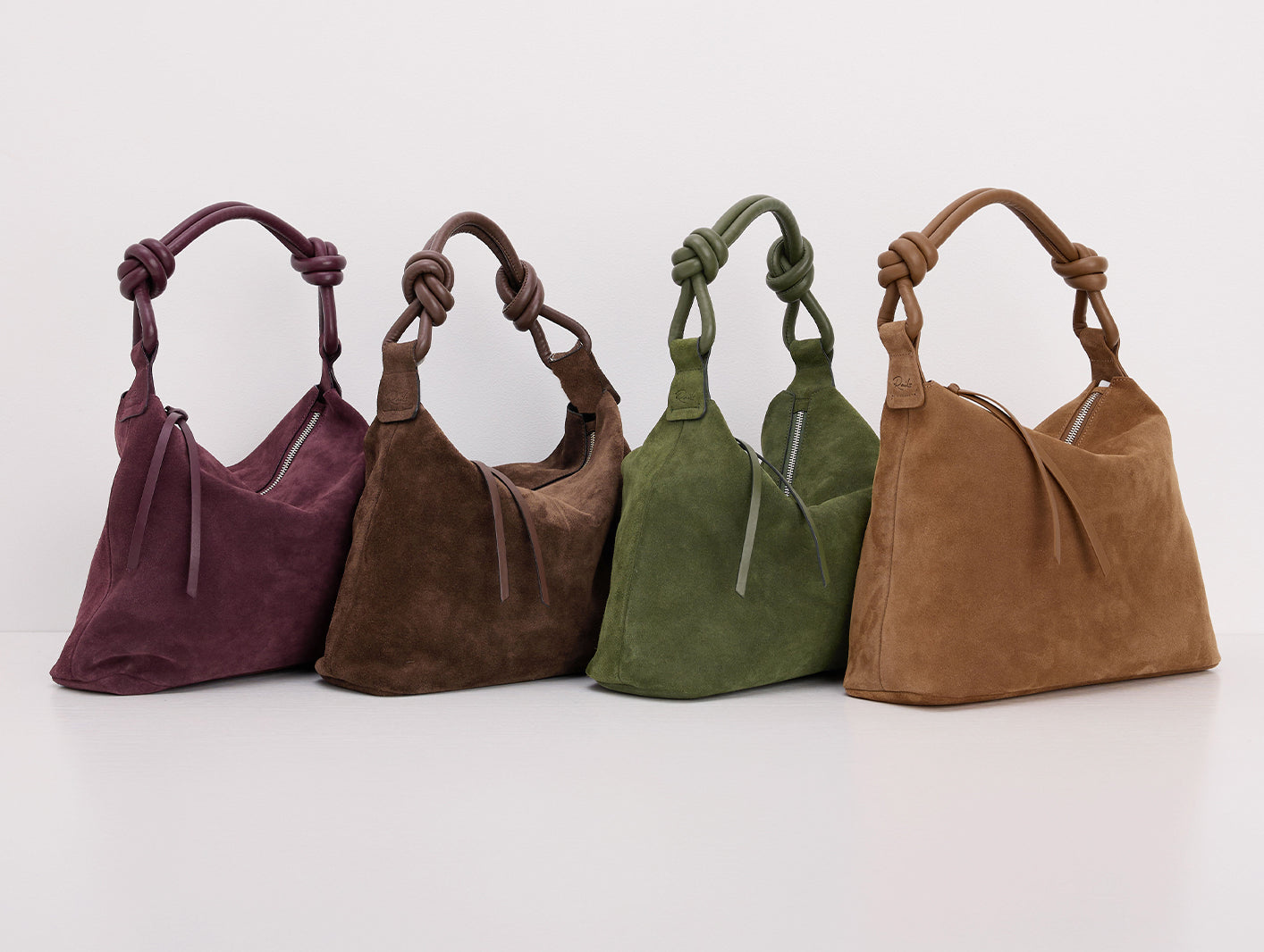 Four suede shoulder bags in plum, taupe, olive green, and camel brown, arranged in a row on a clean white background.