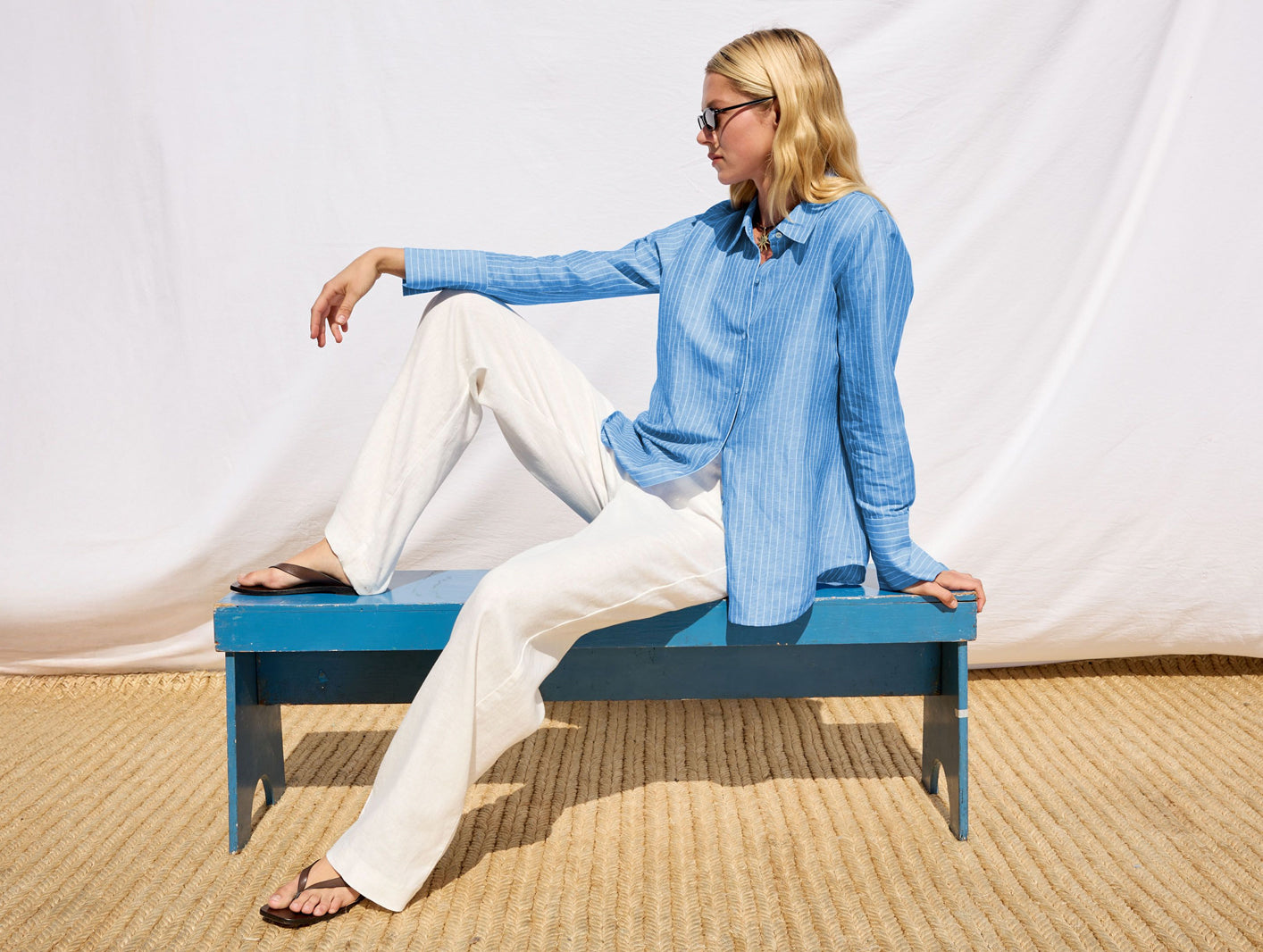Woman wearing a blue linen button down shirt with white linen pants while sitting on a blue bench