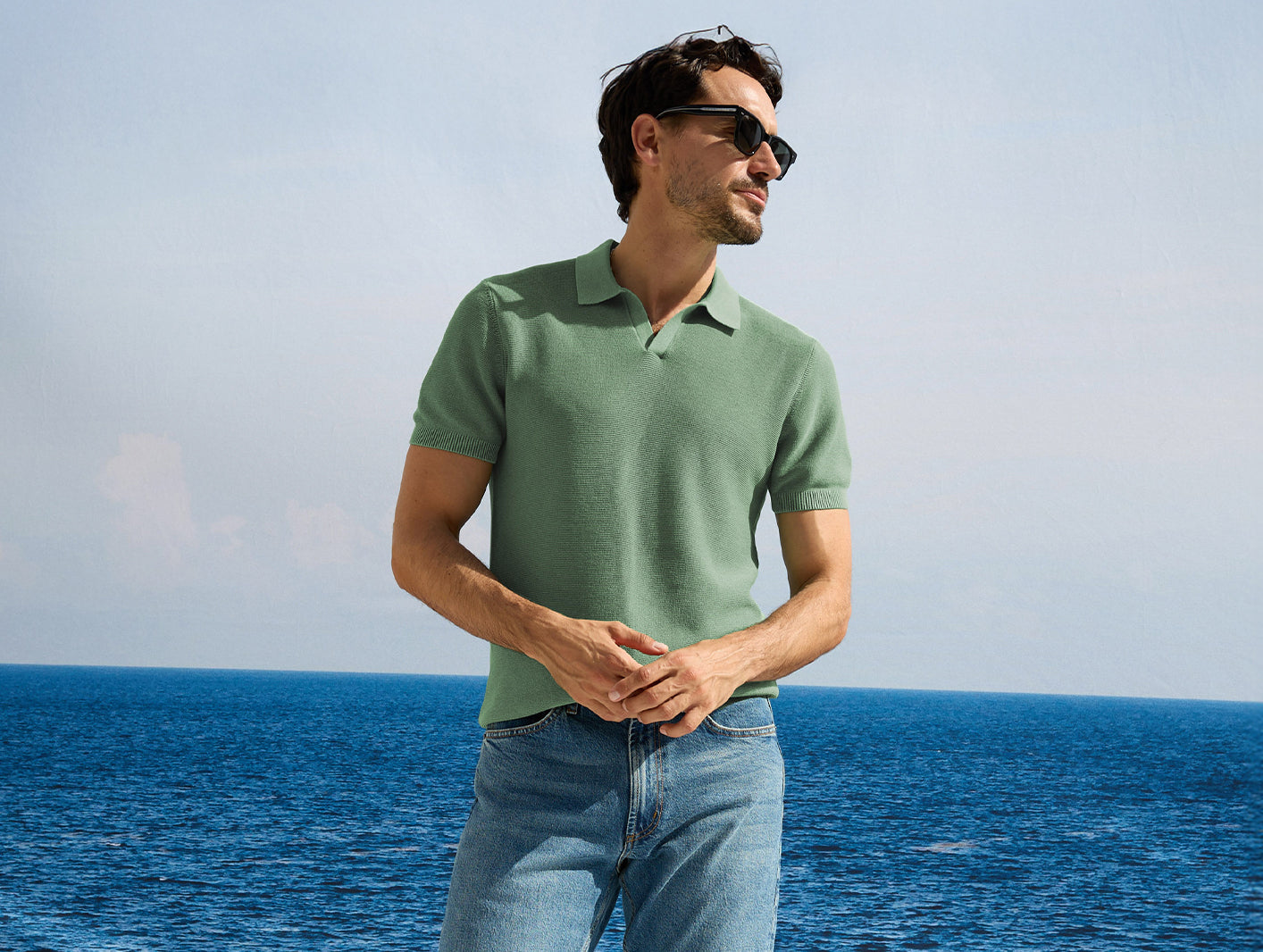 Man wearing a green polo shirt and sunglasses standing by the ocean.