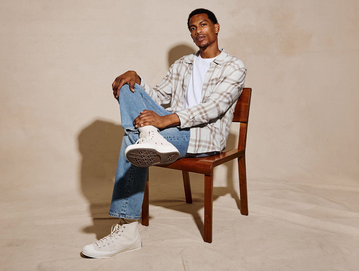 A man sits on a wooden chair wearing a beige and white plaid button-up shirt over a white t-shirt, blue jeans, and white high-top sneakers against a warm beige studio backdrop.