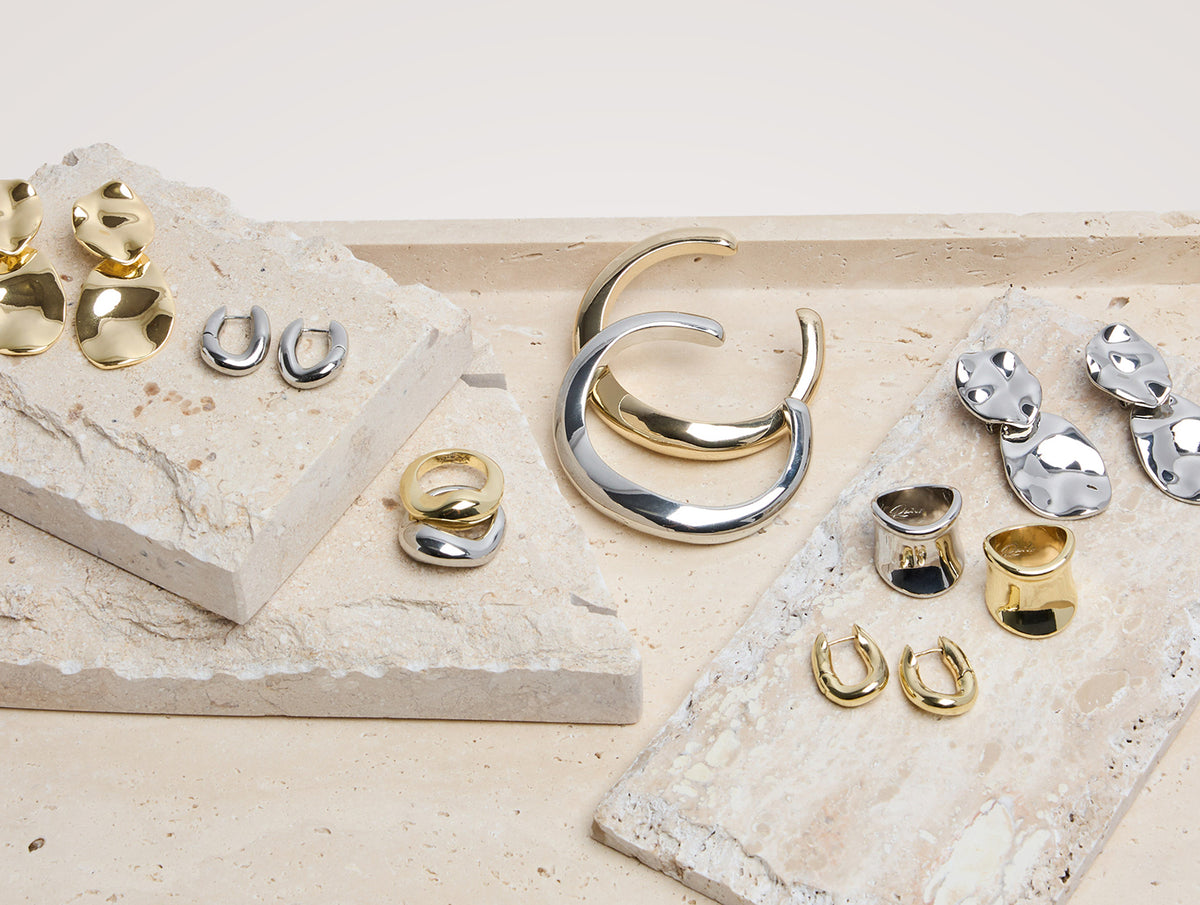 A styled flat-lay of multiple pairs of earrings and rings in polished gold and silver finishes, arranged on textured beige stone slabs against a light neutral background.