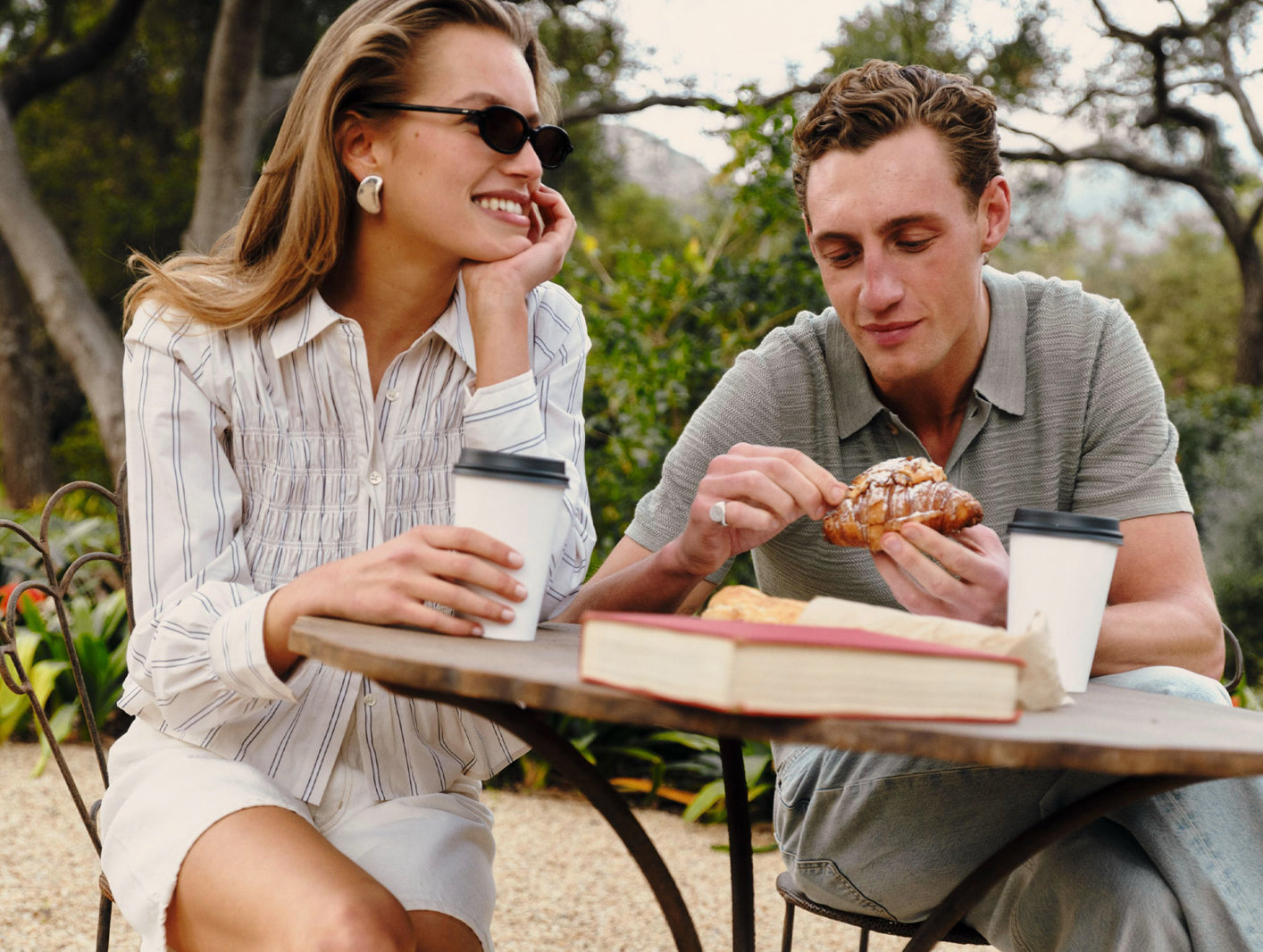 A man and a woman sitting at an outdoor cafe table, sharing coffee and a pastry. The woman is wearing a white button-down shirt with thin blue vertical stripes and white shorts, while the man wears a gray-green striped polo shirt and jeans.