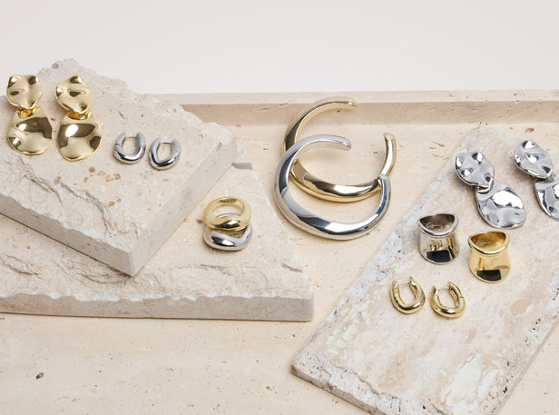 A styled flat-lay of multiple pairs of earrings and rings in polished gold and silver finishes, arranged on textured beige stone slabs against a light neutral background.