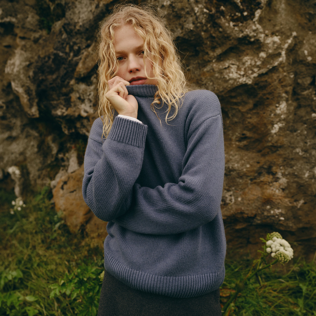 A woman with curly blonde hair wearing a slate blue turtleneck sweater, posing in front of a large rock wall outdoors.