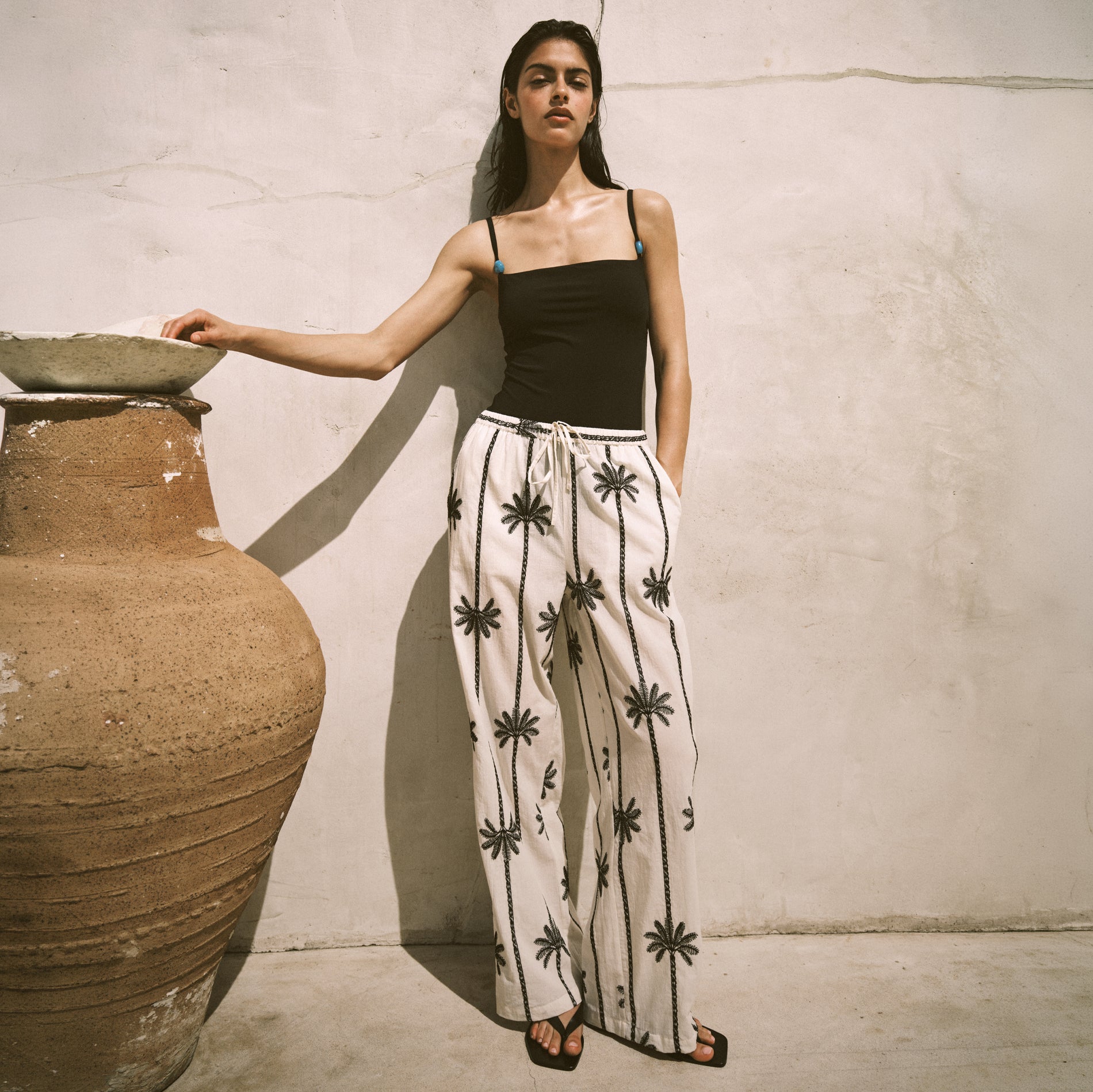 Woman wearing a black top and white pants with black patterns leaning against a large pot.