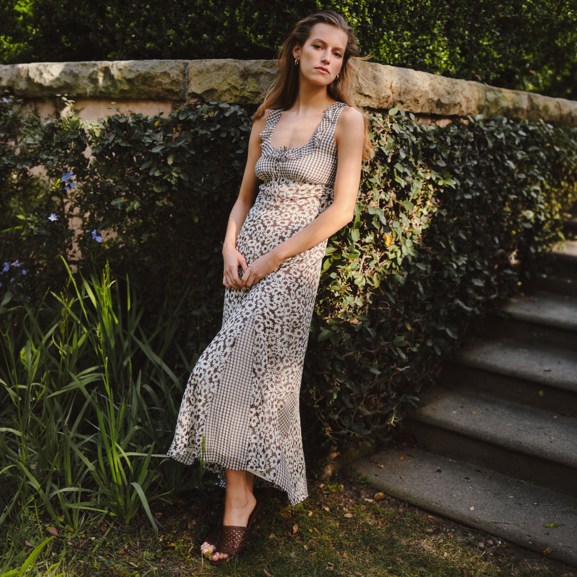 A woman wearing a sleeveless leopard-print maxi dress and brown woven sandals, posing outdoors against a stone wall with green ivy and garden plants.