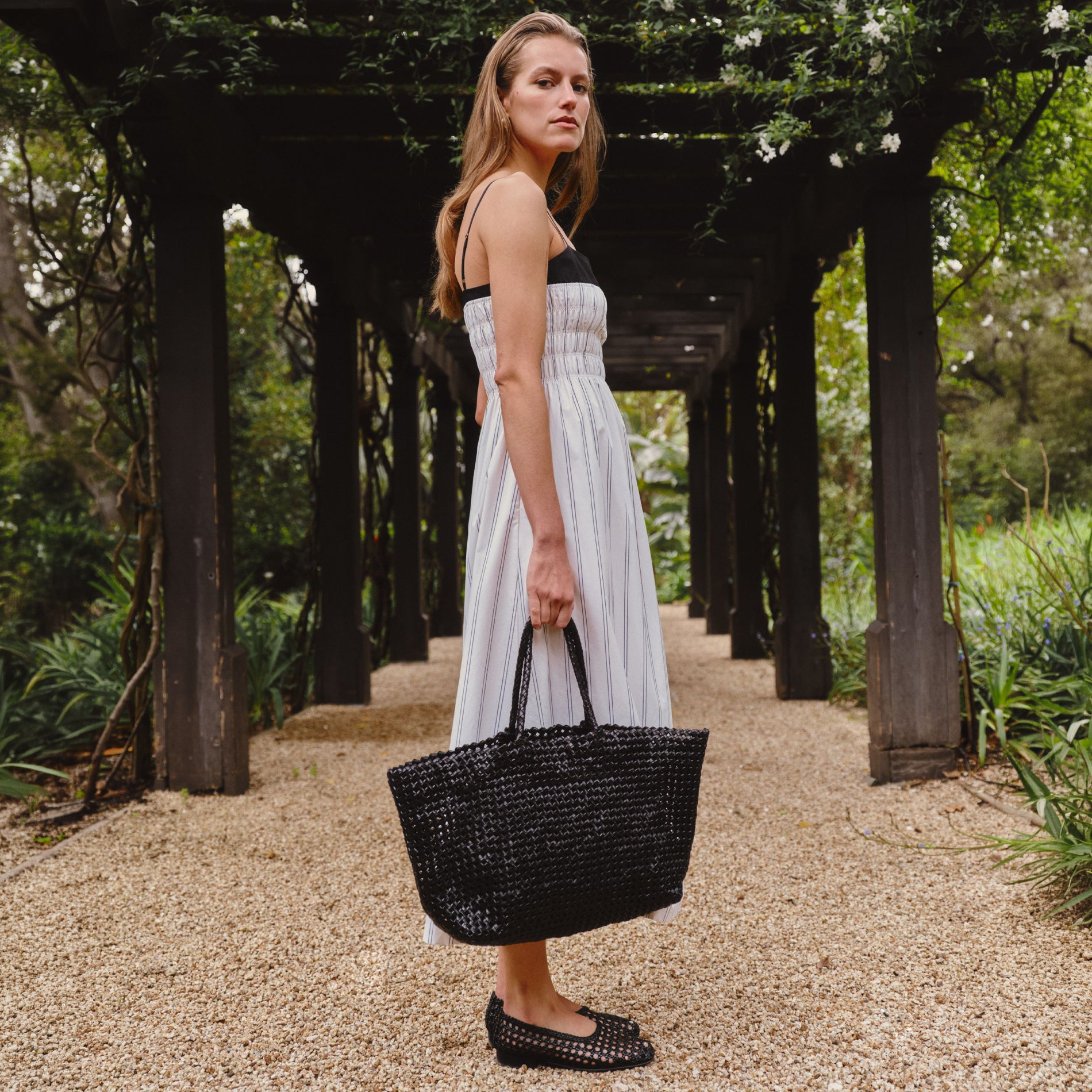 A woman stands on a gravel path under a wooden pergola, wearing a white sleeveless midi dress with a black bodice and holding a large black woven tote bag.