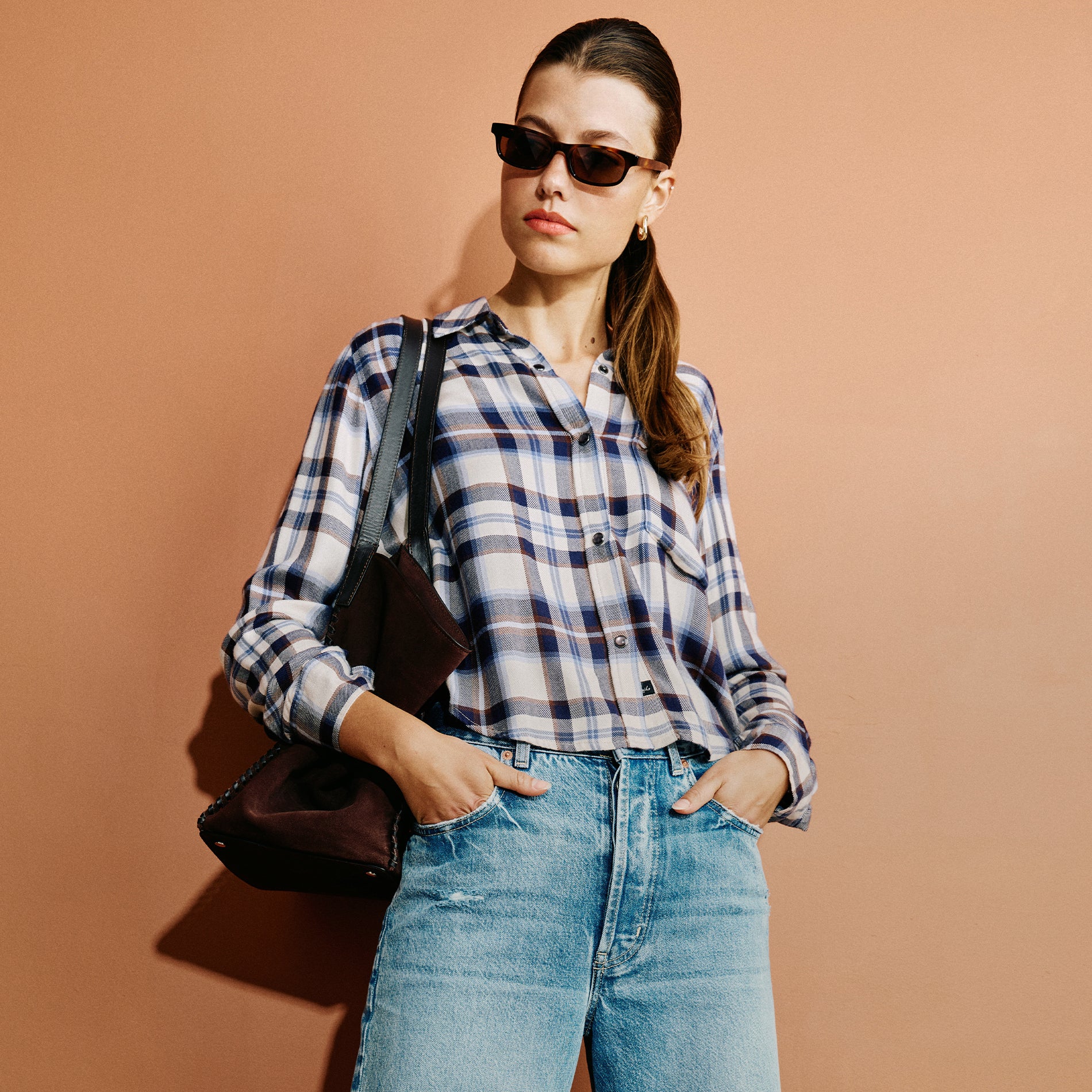 A woman wearing a blue, white, and navy plaid button-up shirt, light-wash blue jeans, and dark sunglasses, carrying a dark brown leather shoulder bag against a solid terracotta background.