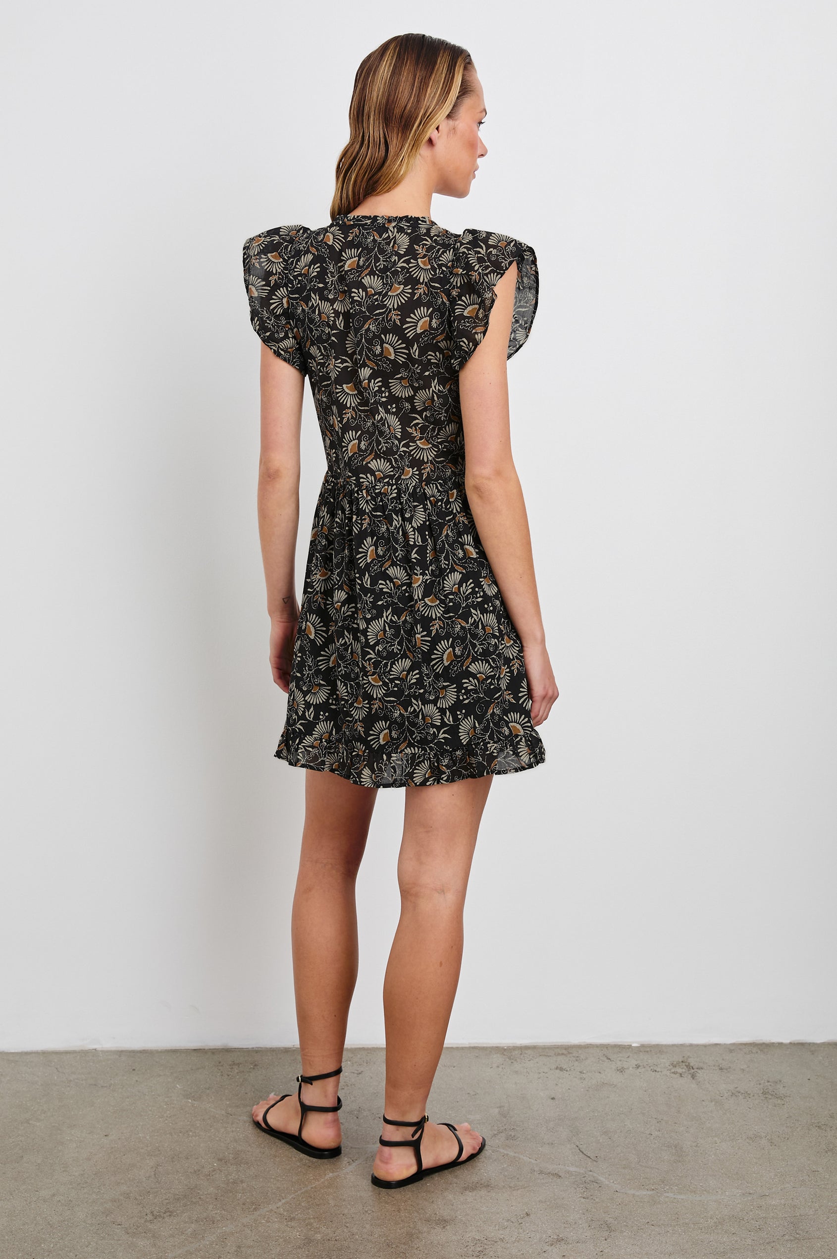 Zinnia Mini Dress with Flutter Cap Sleeves, and a Deep V Neck with Ties in Cassia Colorway, with Orange and Cream Flowers on Black - Back Full Body View Featured on Model 