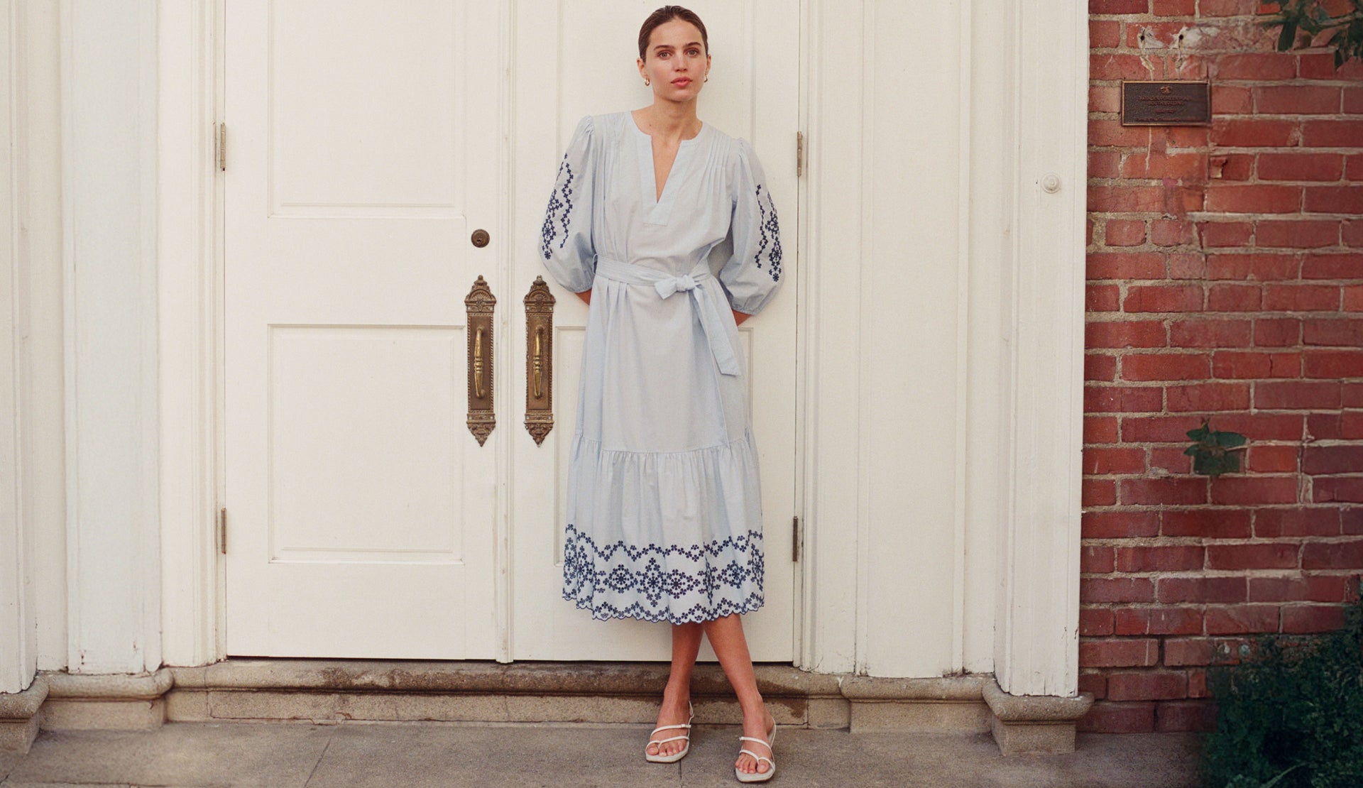 Woman in a light blue dress standing in front of a white door with a red brick wall.