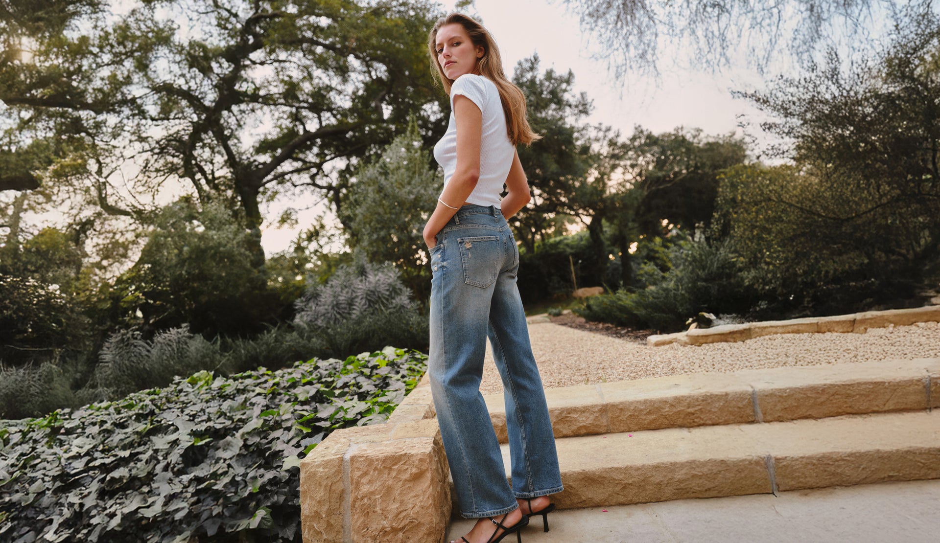 Woman wearing a white top and jeans standing outdoors with trees in the background