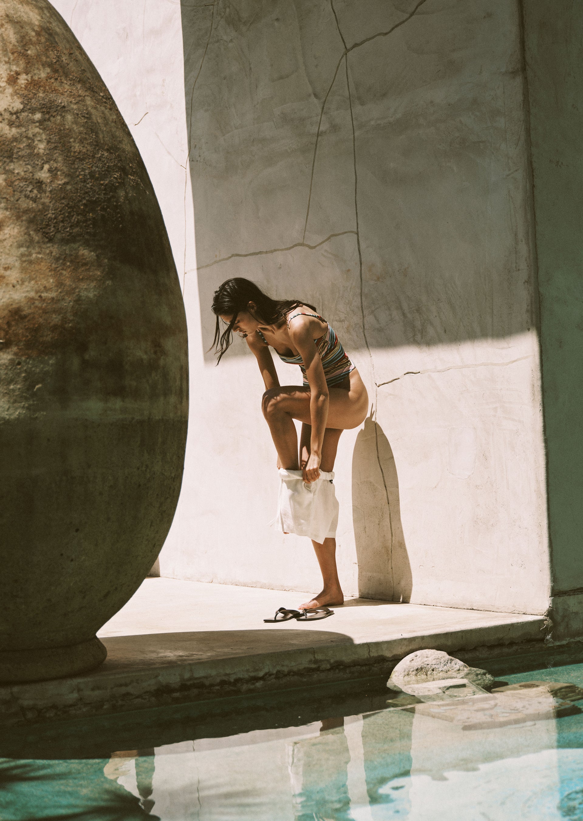 Woman in a swimsuit standing by a pool with large stones and concrete walls.