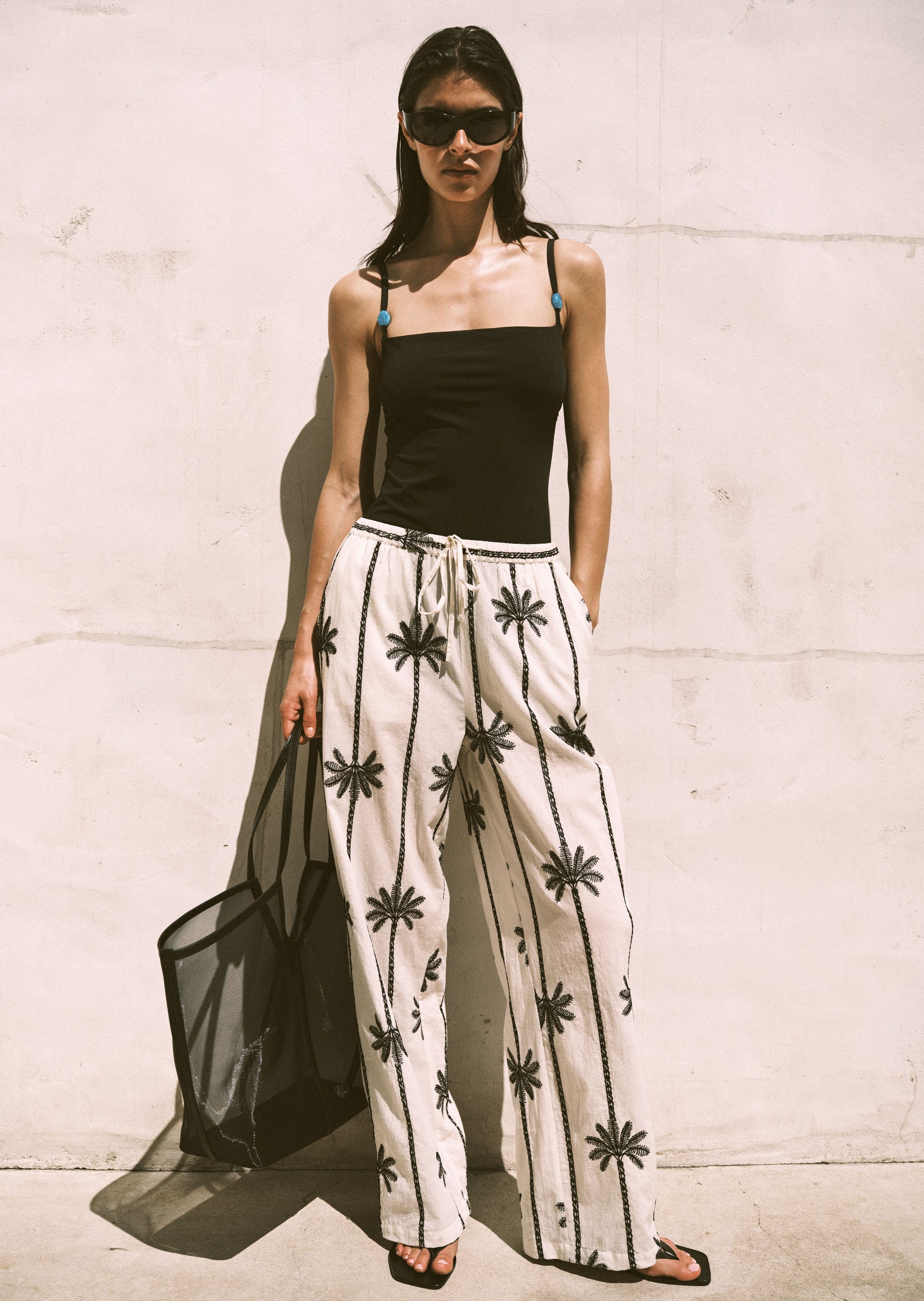 Woman wearing a black top and beige pants with black patterns against a light-colored wall.